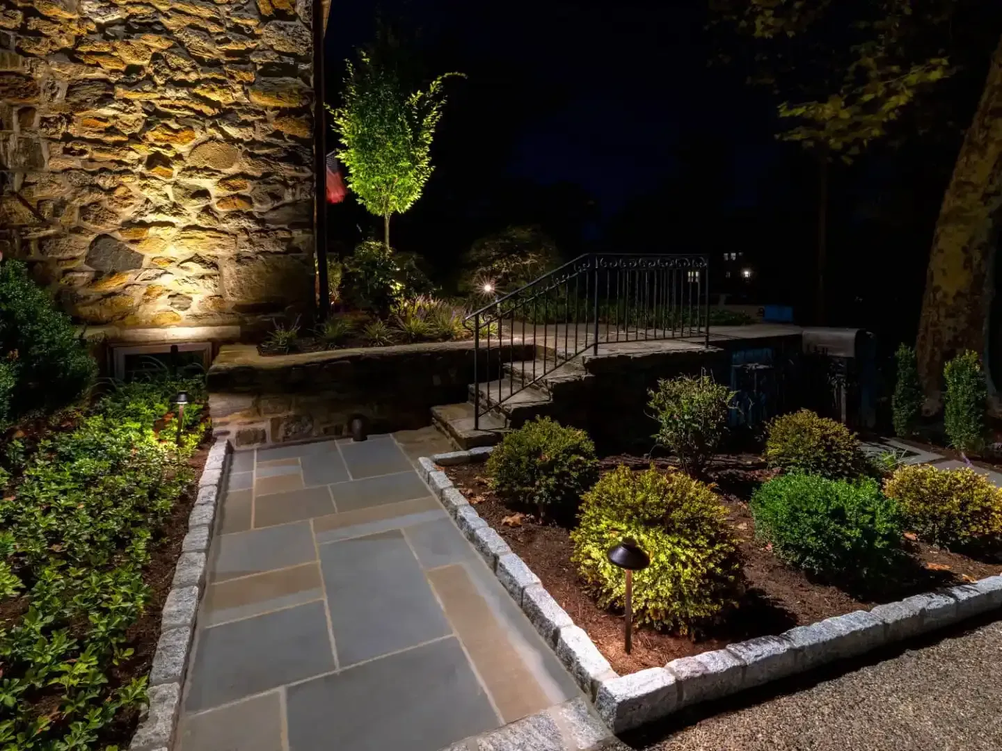 Stone walkway through a lit landscaped garden at night, with trimmed shrubs and stone walls