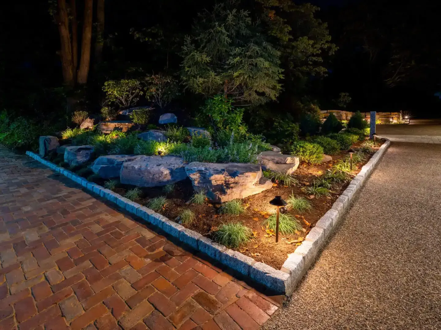 Lit landscaped garden bed with rocks and shrubs beside a brick walkway at night