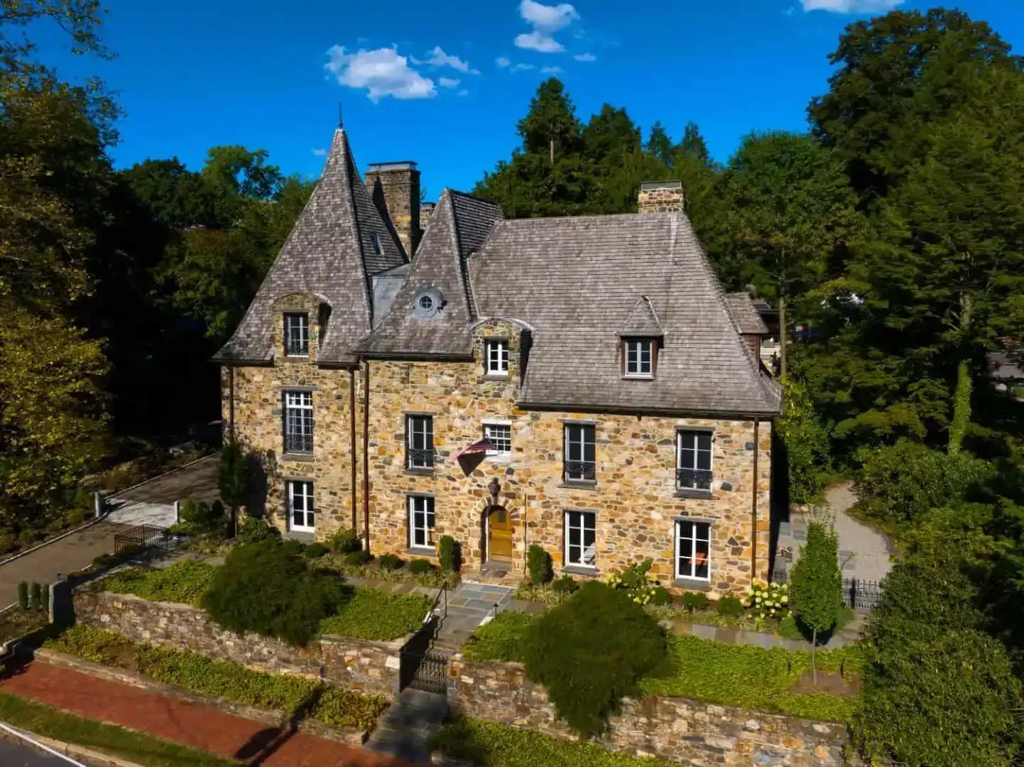 Stone manor house with steep slate roof and dormers, surrounded by trees under a bright blue sky