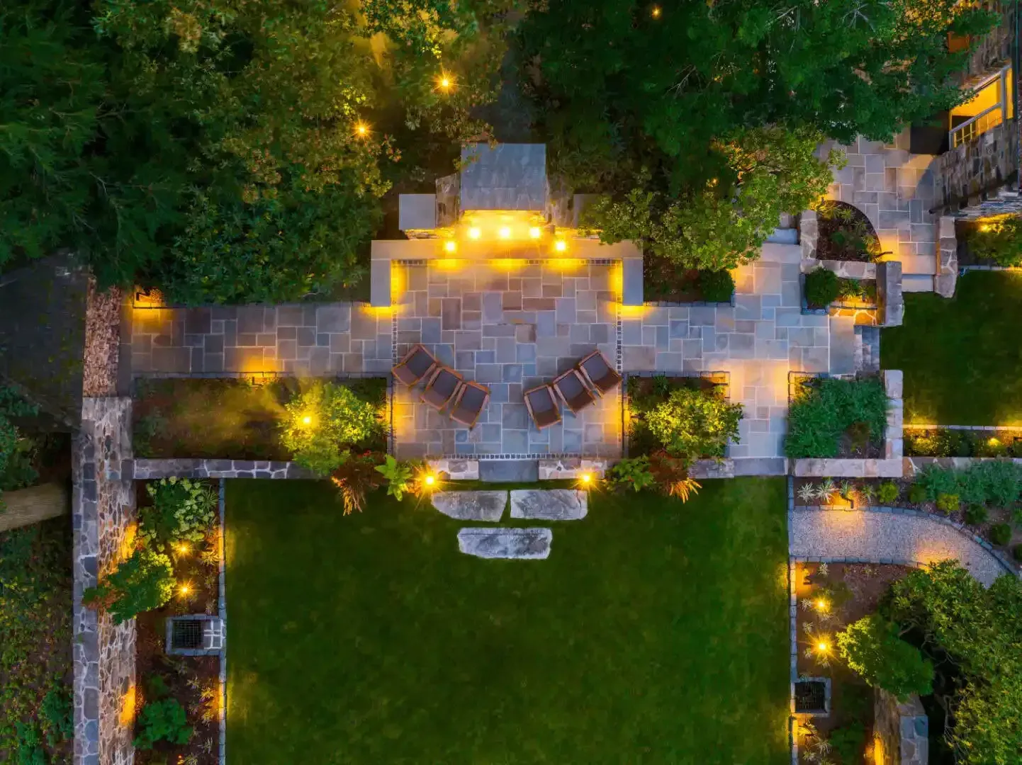 Aerial view of a lit stone patio and walkway surrounded by lush green trees at dusk.