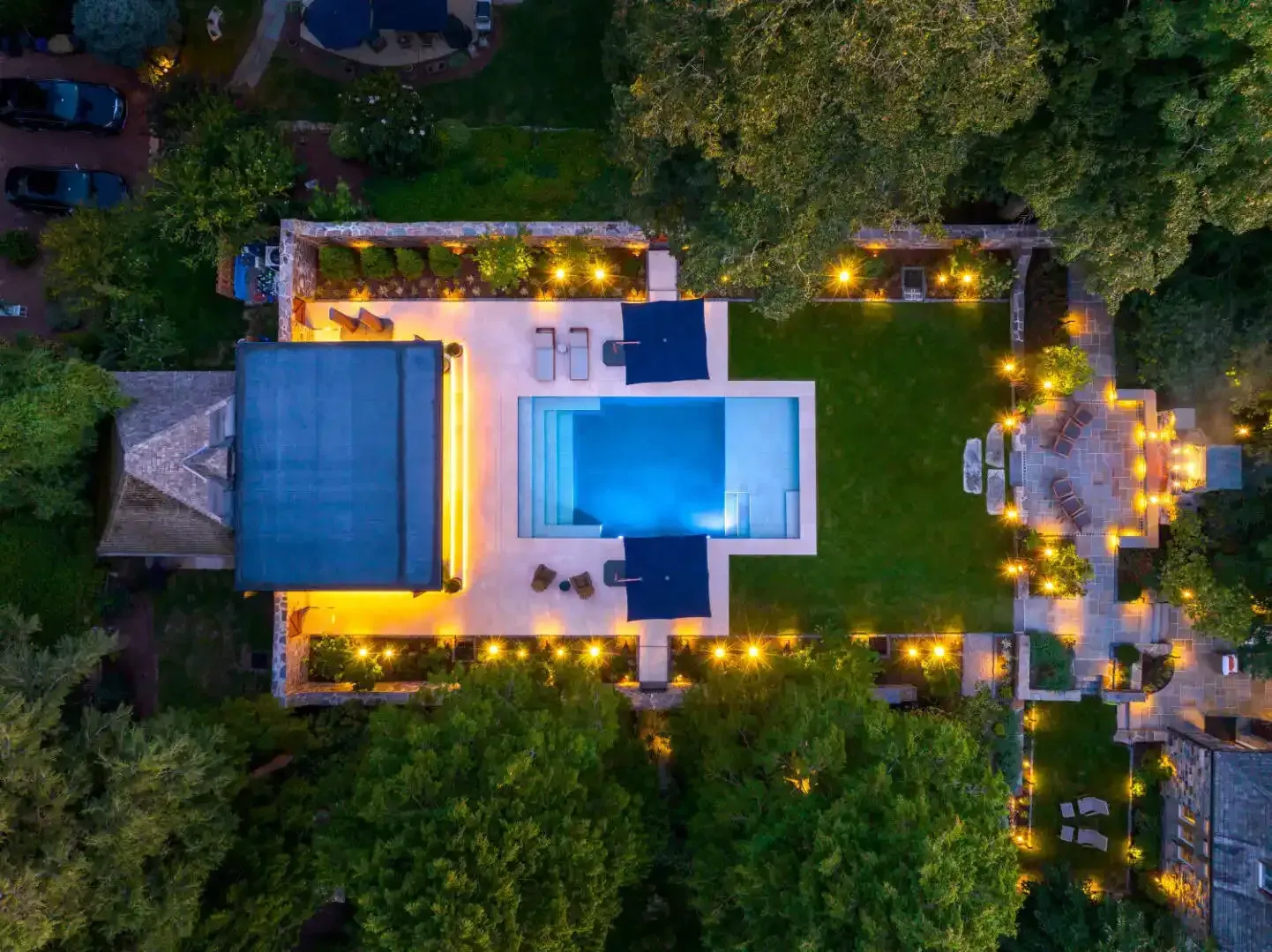 Aerial view of a backyard with a lit swimming pool, patio, and surrounding trees at dusk