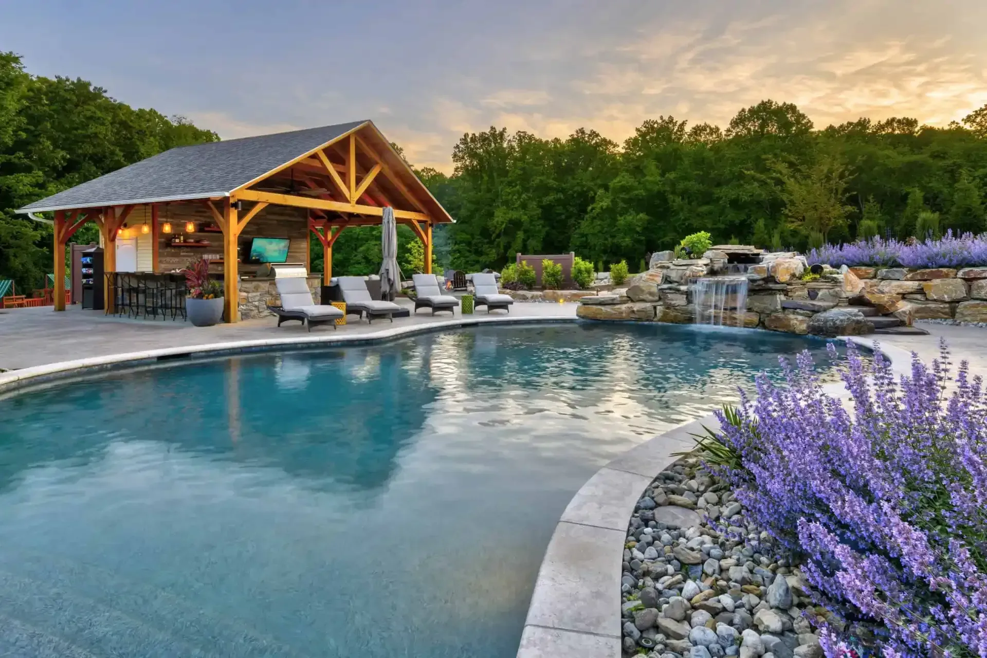 Luxury backyard pool with cabana, lounge chairs, stone waterfall, and purple flowers at sunset