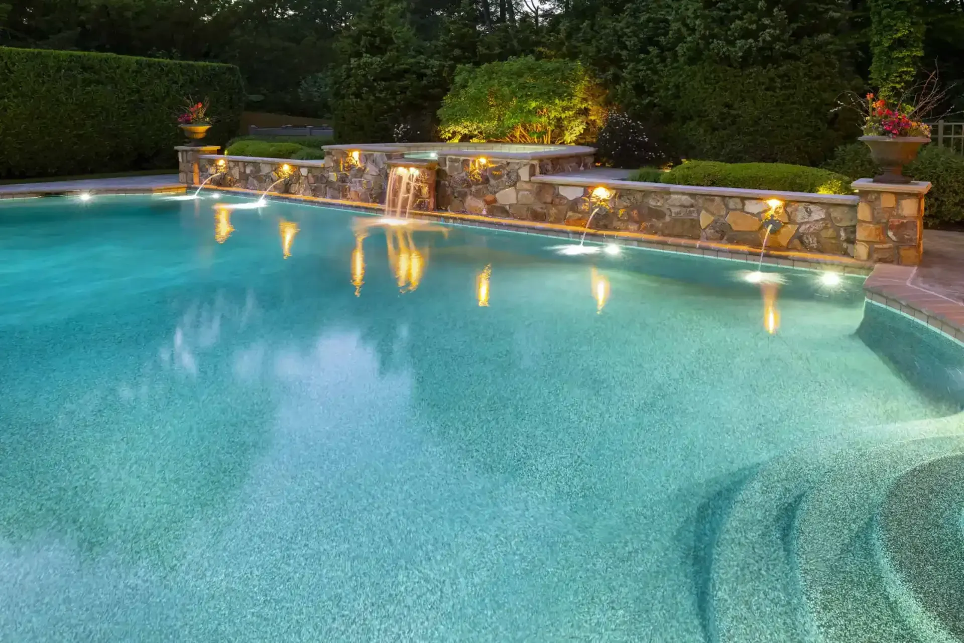 Turquoise swimming pool with a stone waterfall wall and soft underwater lights at dusk