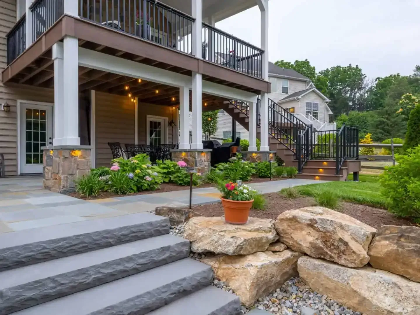 Backyard deck with white columns, stone steps, potted plants, and a landscaped garden beside a house