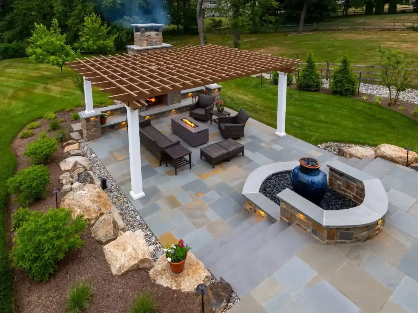 Outdoor patio with pergola, seating, fire pit, and stone landscaping in a green backyard.