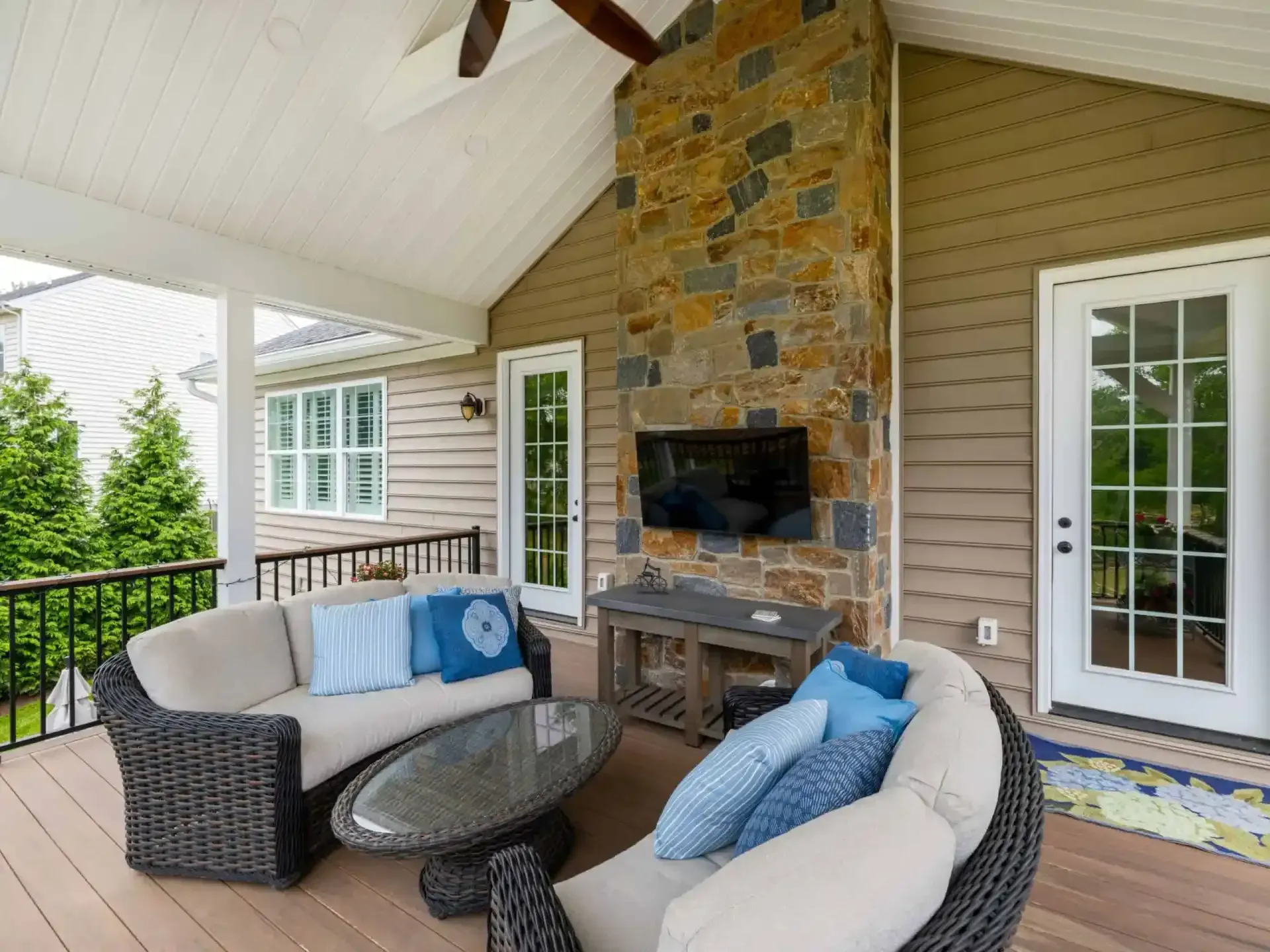Covered patio with wicker seating, glass table, TV, and stone fireplace next to a home exterior