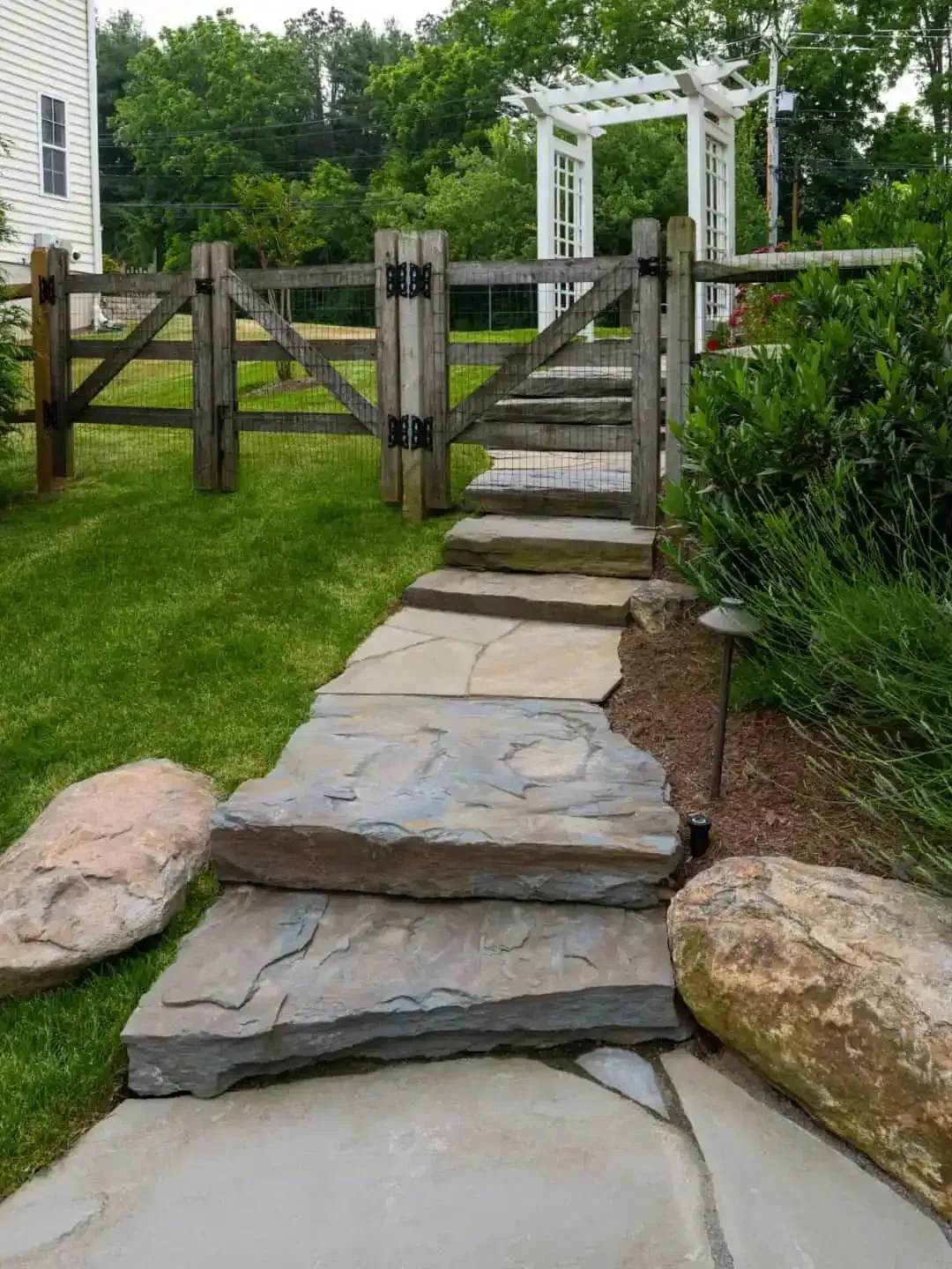 Stone path and steps leading to a wooden garden gate and white arbor in a landscaped yard.