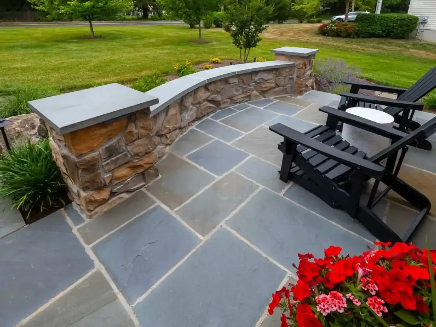 Stone patio with black chairs, low stone wall, and red flowers beside a green lawn.