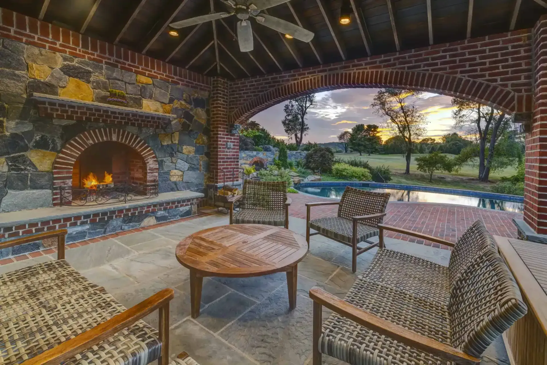 Stone patio with arched fireplace, woven chairs, fire pit, and sunset view over a landscaped yard