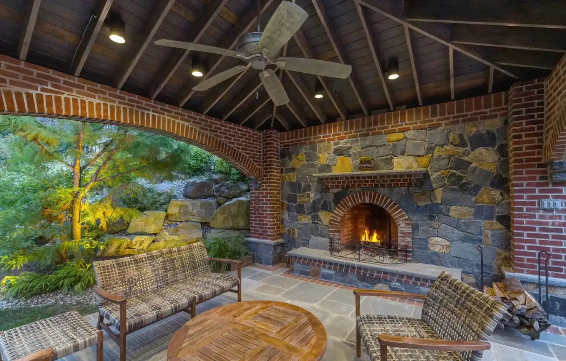 Covered patio with stone fireplace, wicker chairs, and a view of green trees outside.