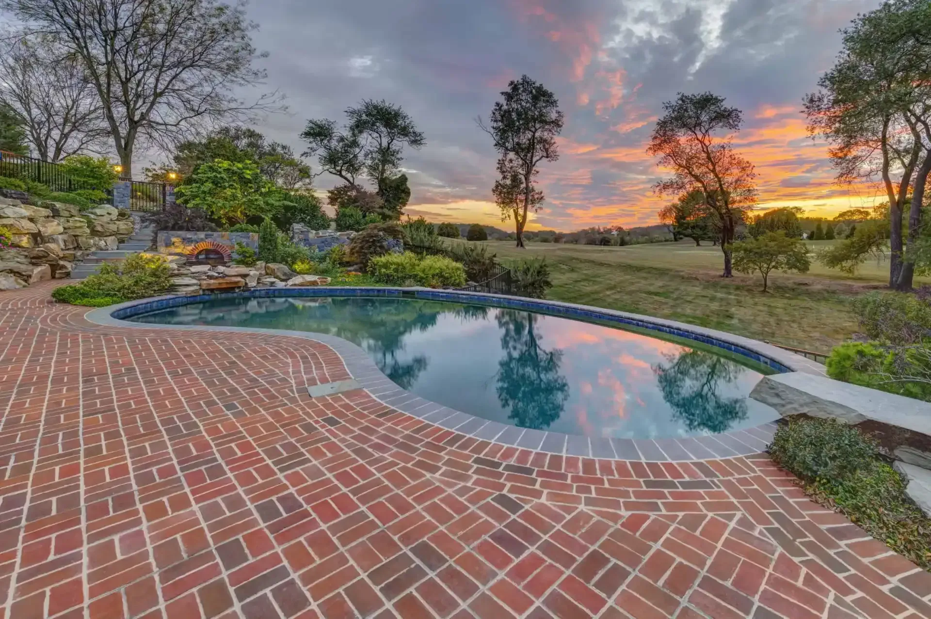 Brick patio with a small backyard pool at sunset, surrounded by trees and a landscaped garden.