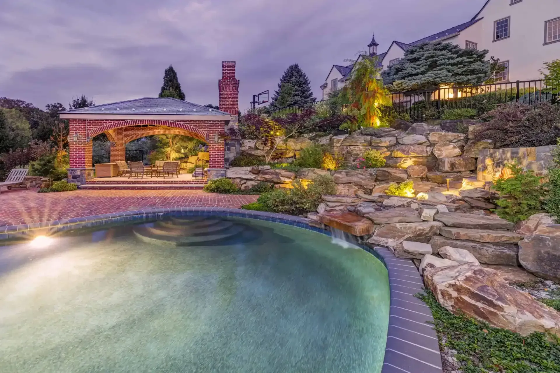 Curved backyard pool beside stone patio, lit waterfall grotto, and hillside landscaping at dusk