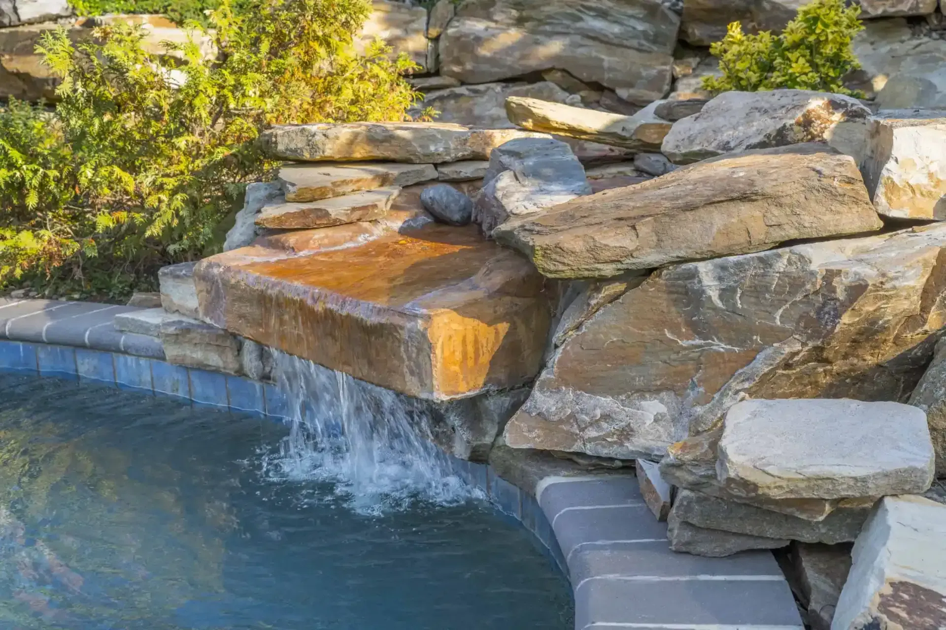 Rock waterfall spilling into a backyard pool with stacked stone landscaping and green shrubs