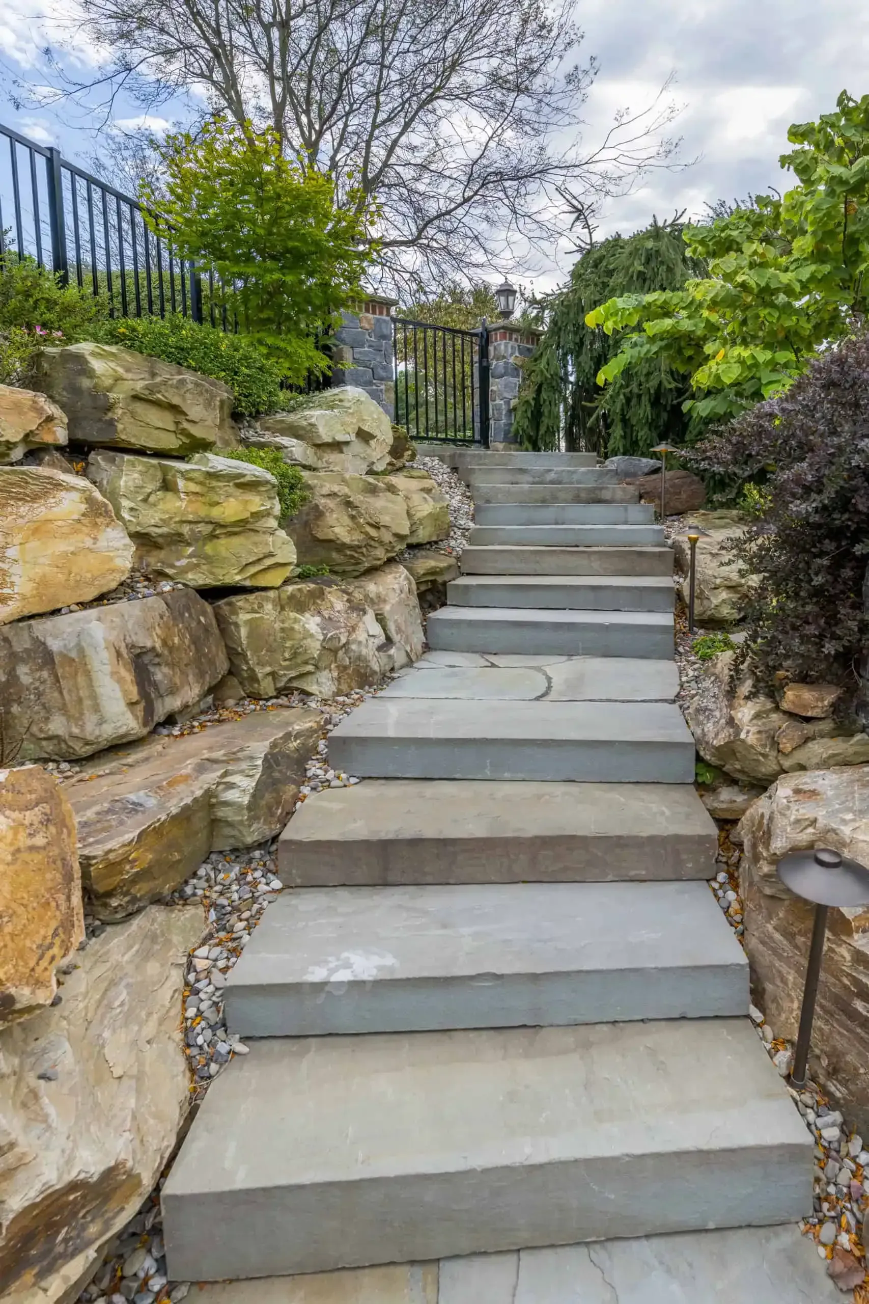Stone garden steps between rock walls leading uphill to a fence and trees.