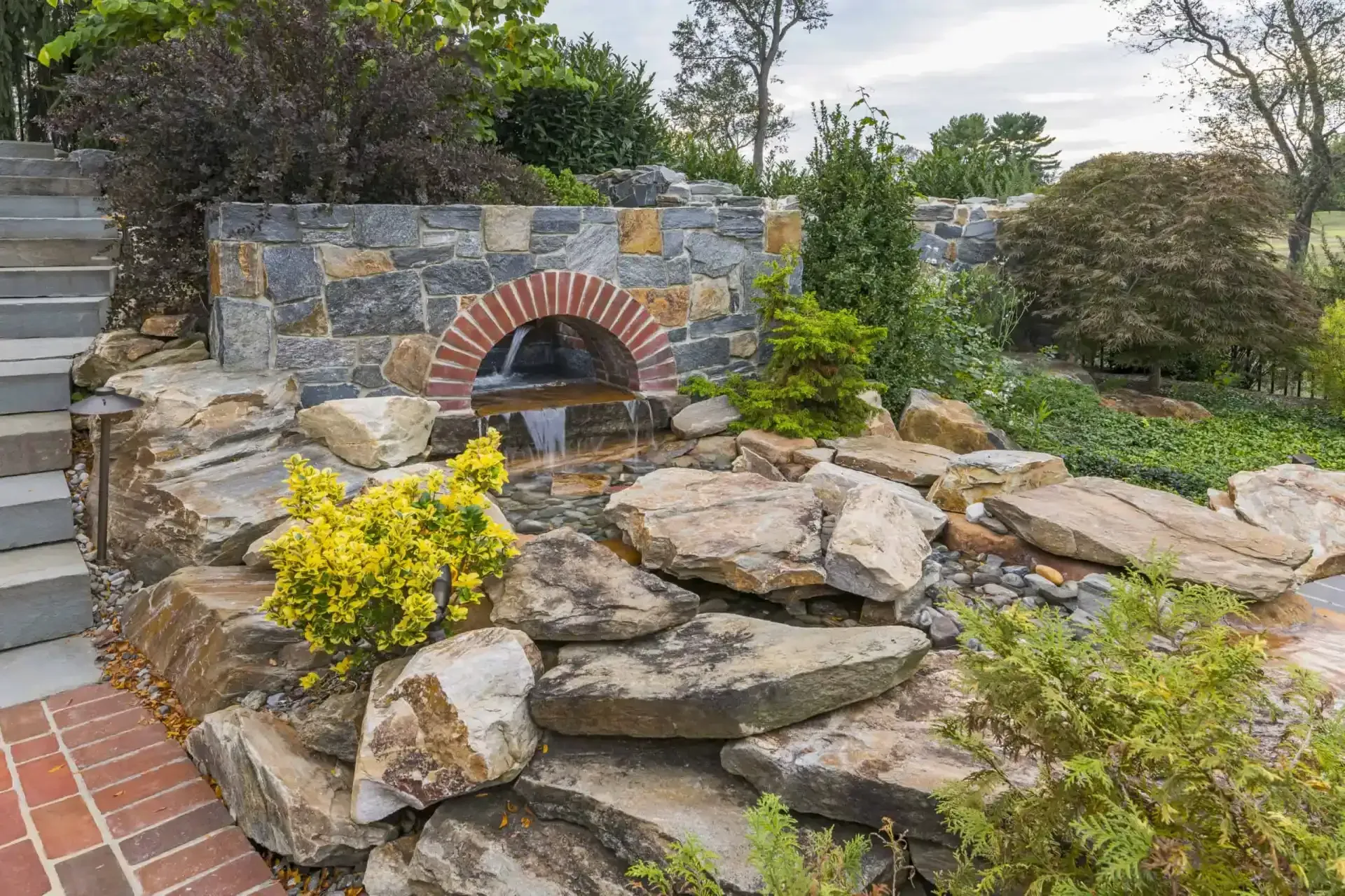 Stone outdoor oven in a landscaped garden with stacked rocks, yellow flowers, and greenery