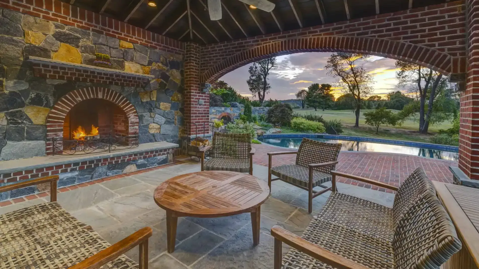 Covered patio with brick fireplace, wicker chairs, and a pool view at sunset