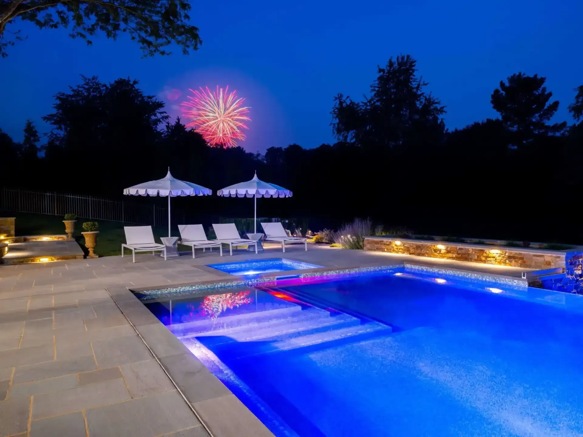 Illuminated backyard pool at night with lounge chairs, umbrellas, and fireworks in the sky
