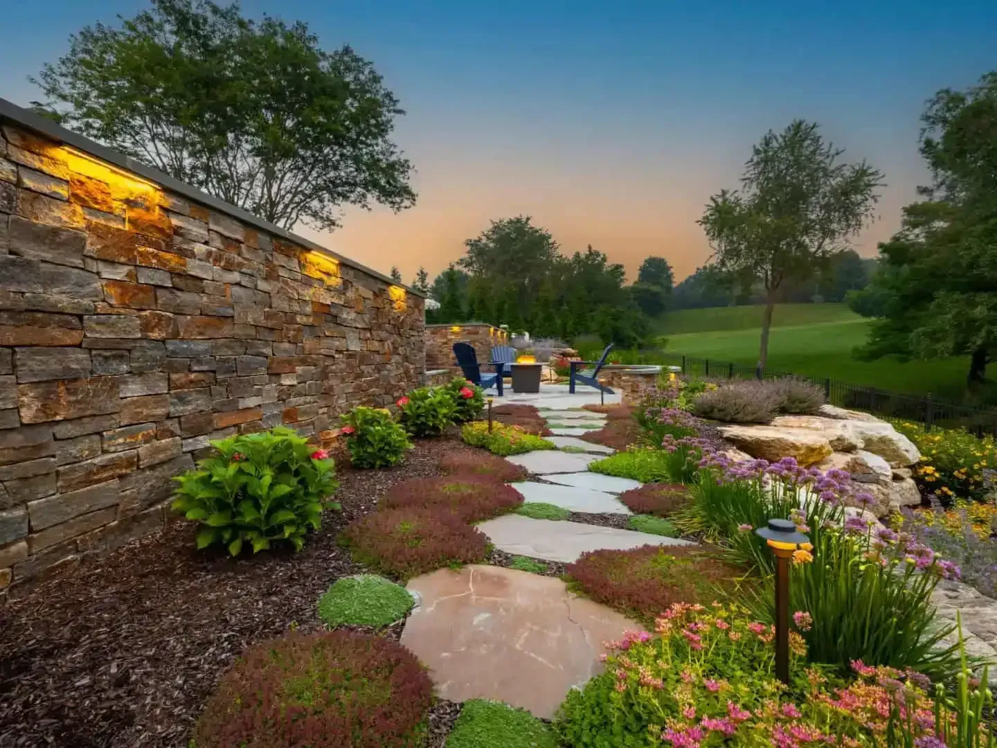 Stone garden path beside a lit retaining wall, with flowers, shrubs, and a sunset sky