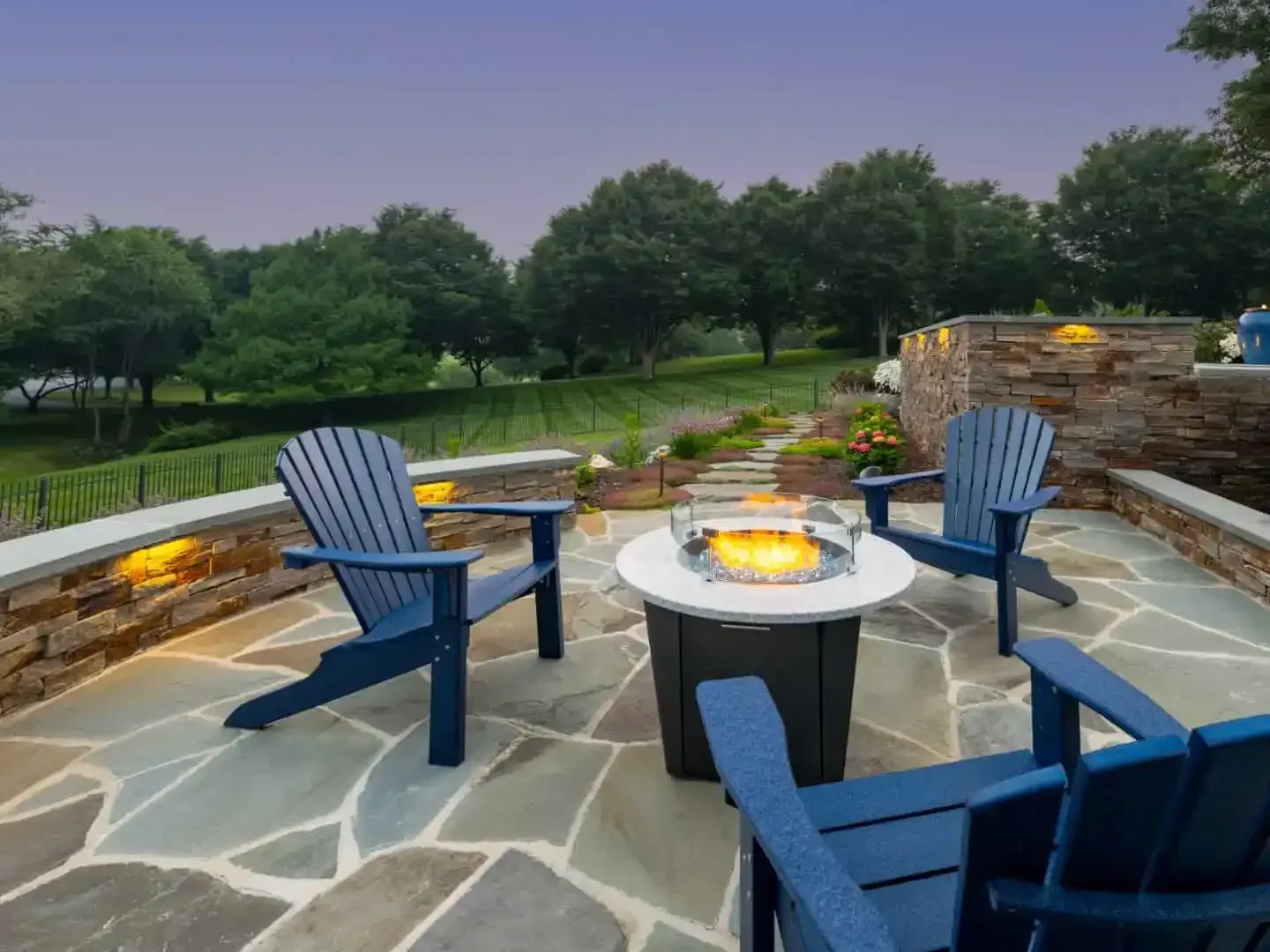 Blue chairs around a stone fire pit on a patio overlooking a green lawn at dusk