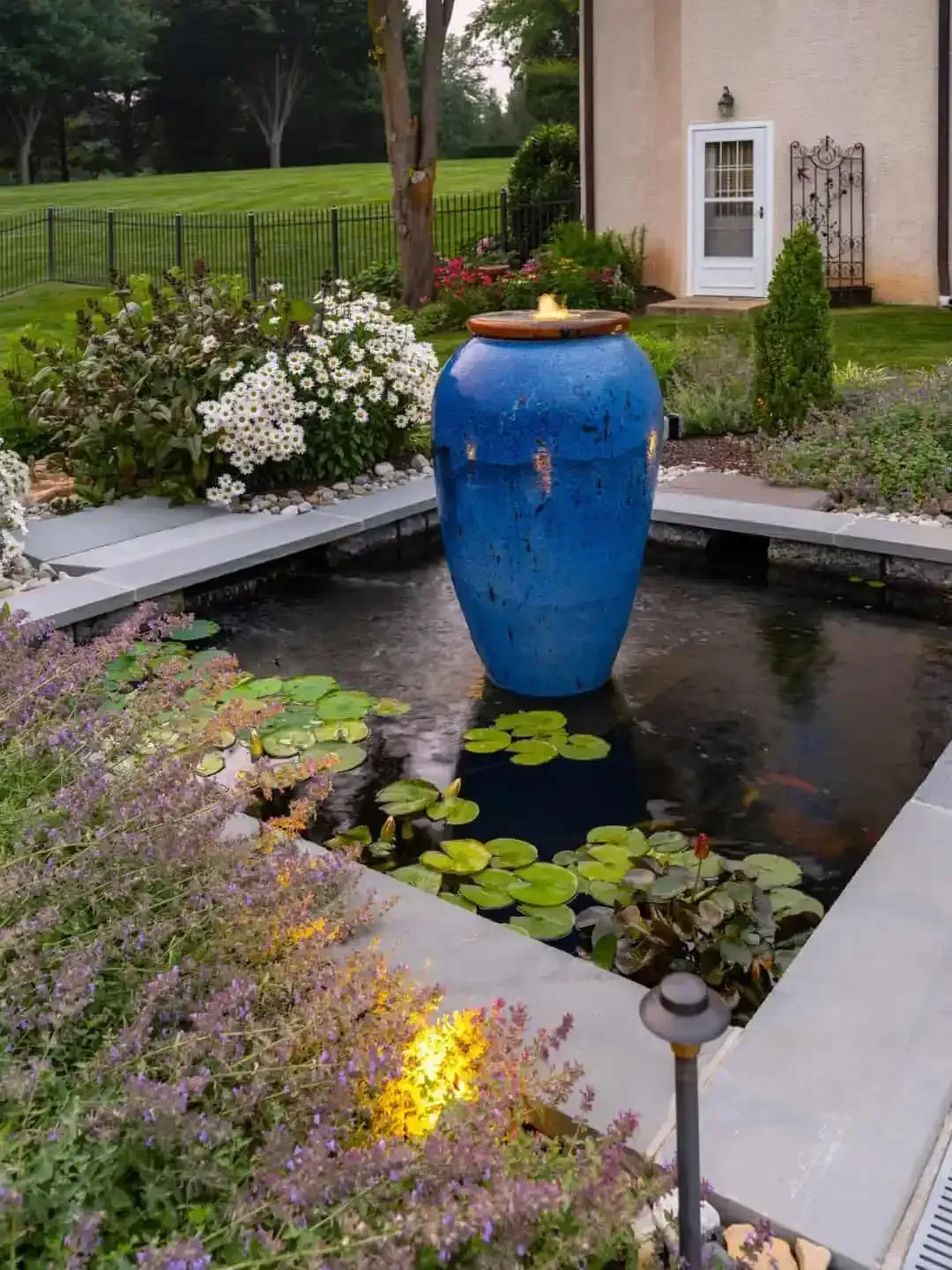 Blue ceramic urn fountain in a garden pond surrounded by white flowers and greenery