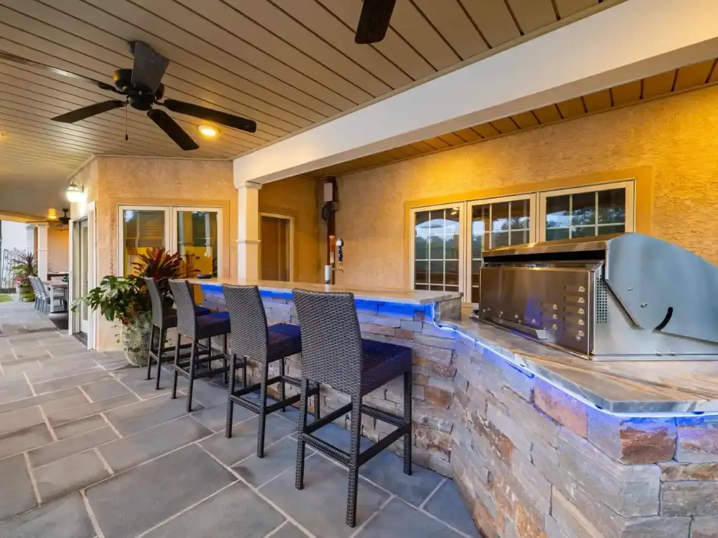 Covered patio with stone bar, blue accent lighting, stools, and ceiling fans at night