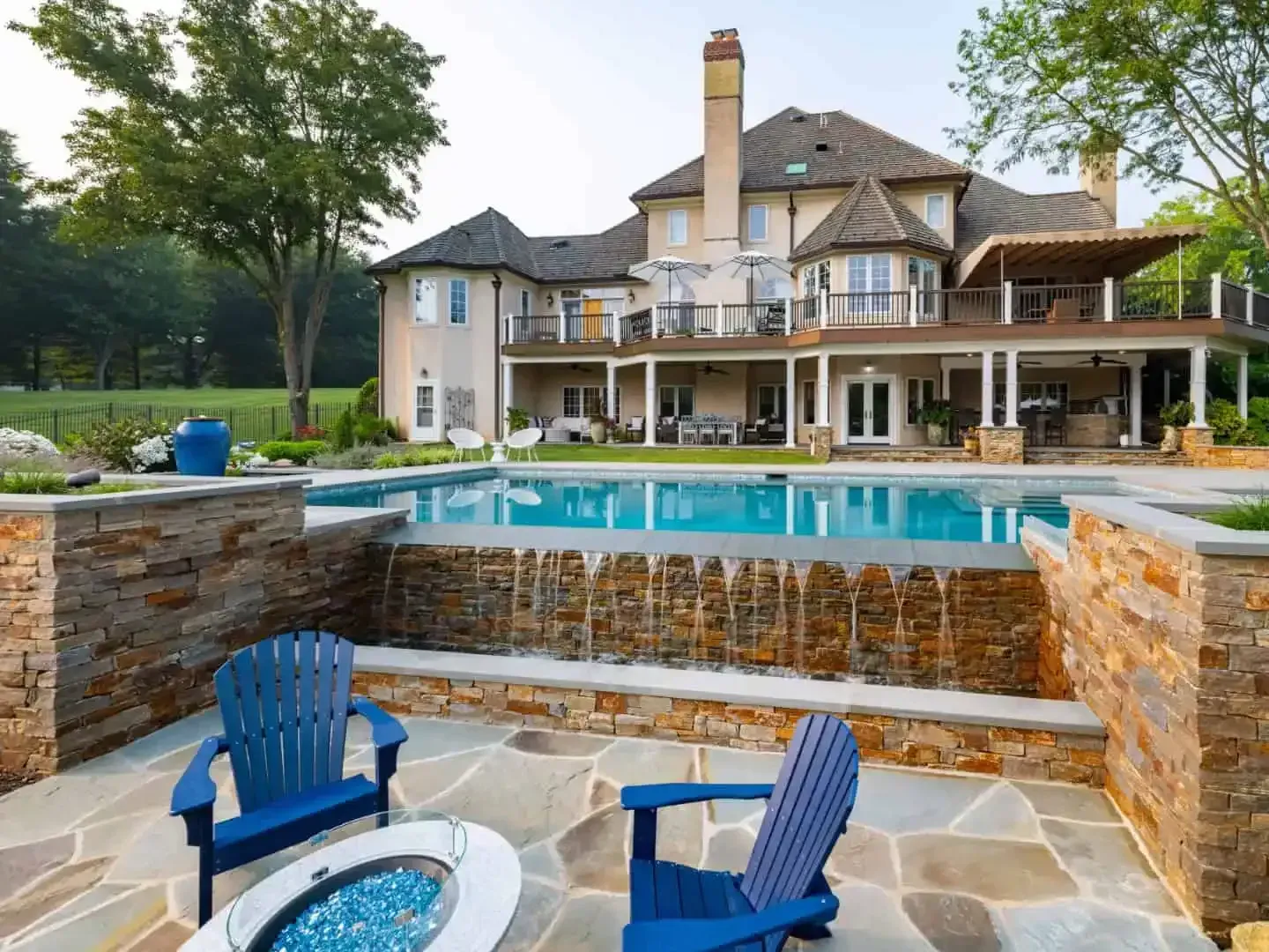 Luxury house with a pool, stone patio, and blue Adirondack chairs at sunset