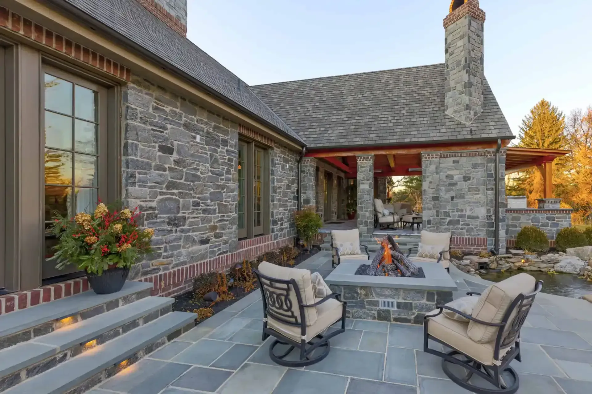 Stone patio with outdoor fire pit, cushioned chairs, and a rustic stone house in the background