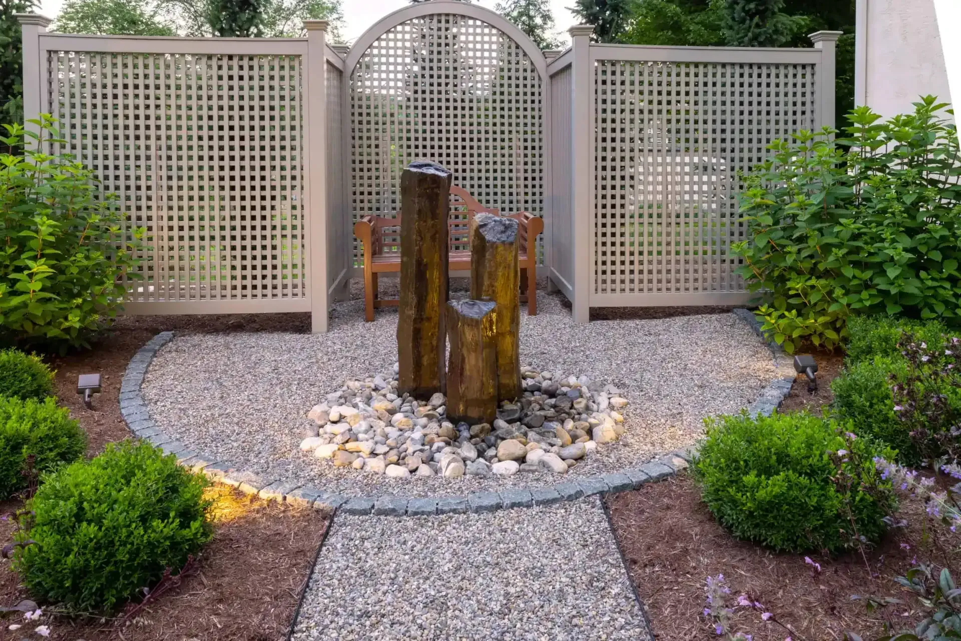Courtyard fountain with wooden sculpture, gravel path, and lattice garden screens bordered by shrubs