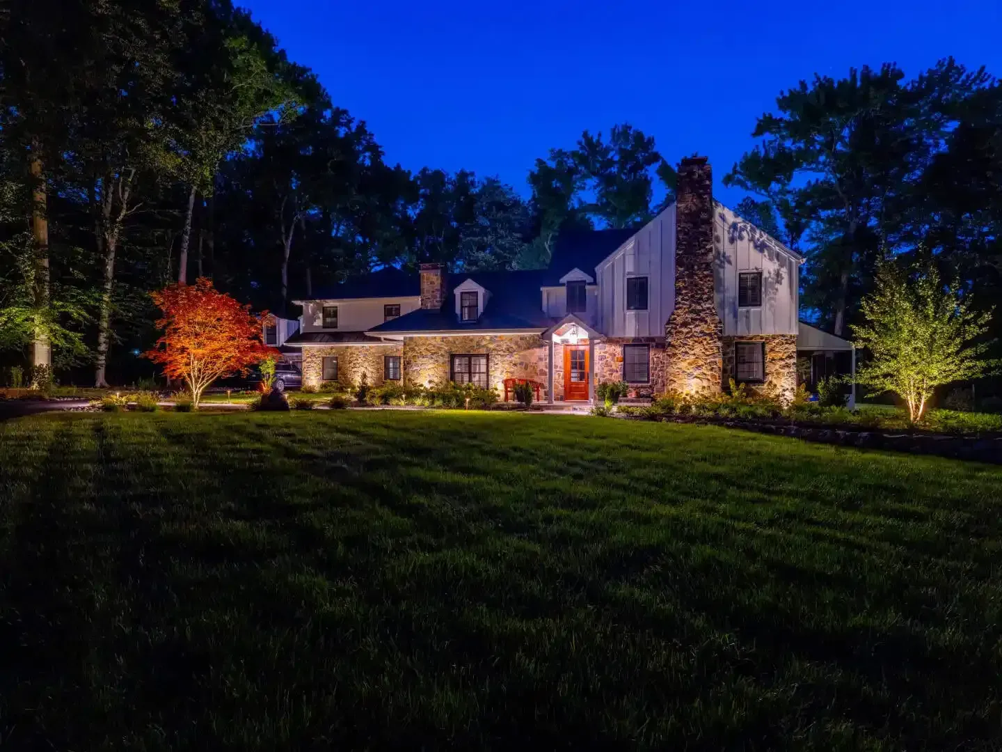 Large stone house lit at night, with warm porch lights and illuminated trees under a deep blue sky.