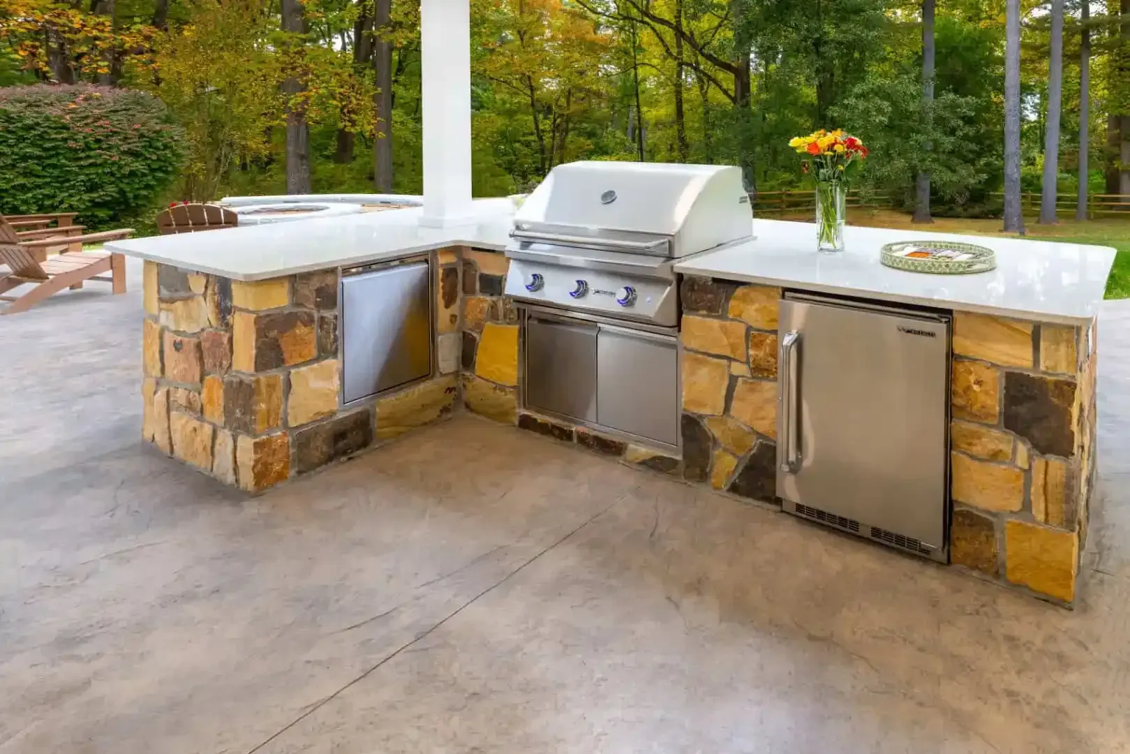 Outdoor stone patio kitchen with grill and stainless steel cabinets, backed by trees and greenery