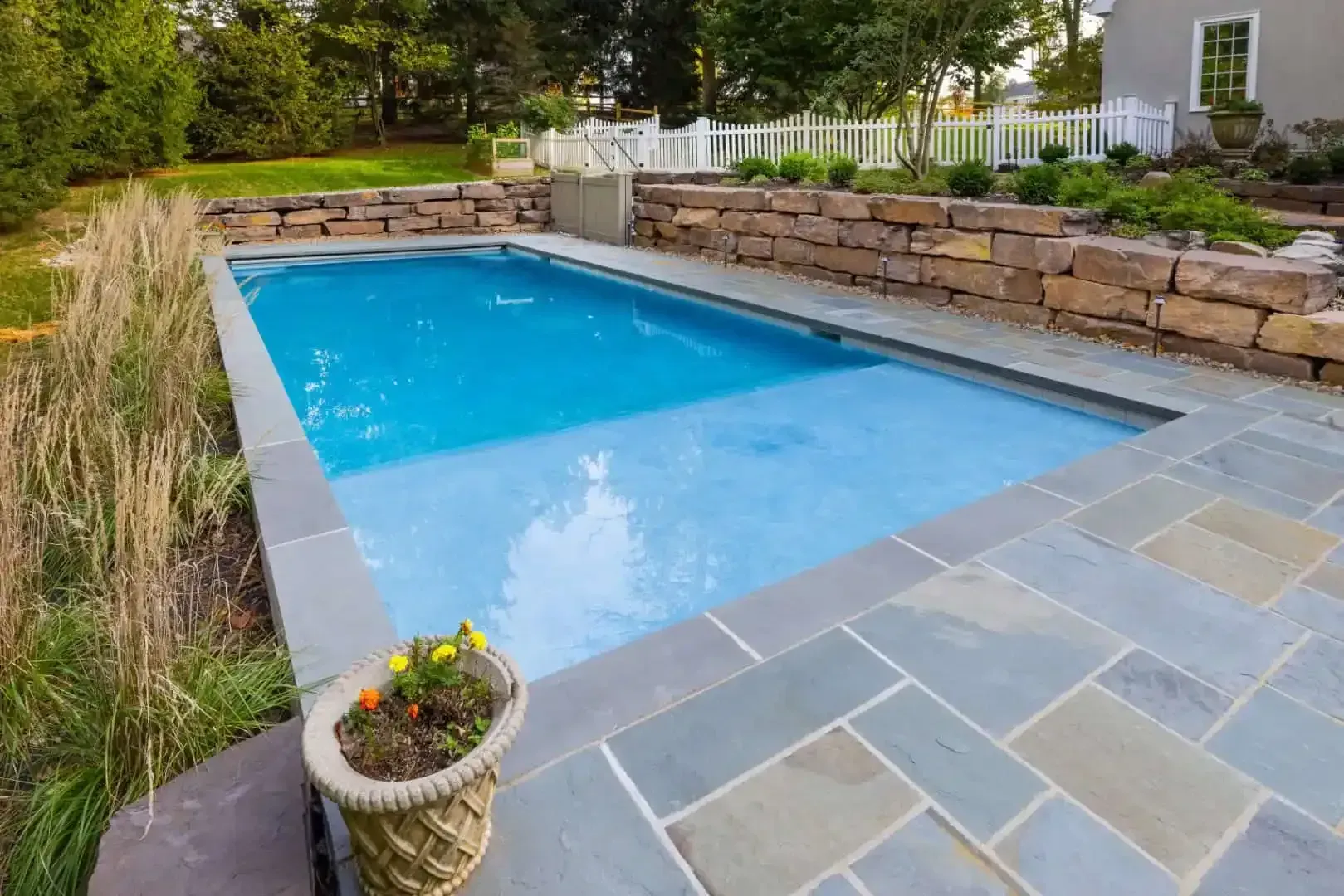 Backyard swimming pool with blue water, stone patio, and low stone retaining wall surrounded by greenery