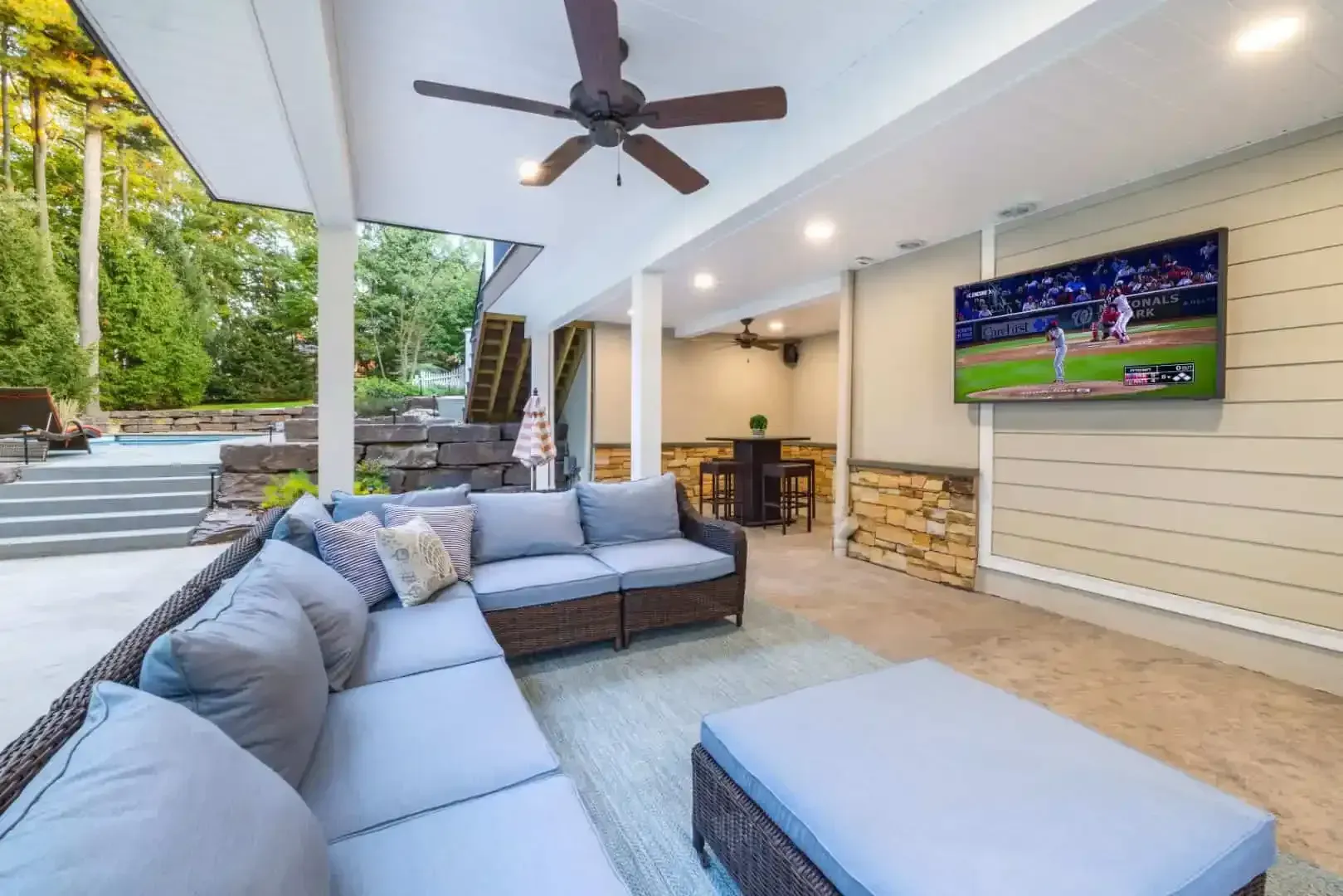 Covered patio lounge with gray sectional sofa, ceiling fan, and wall-mounted TV
