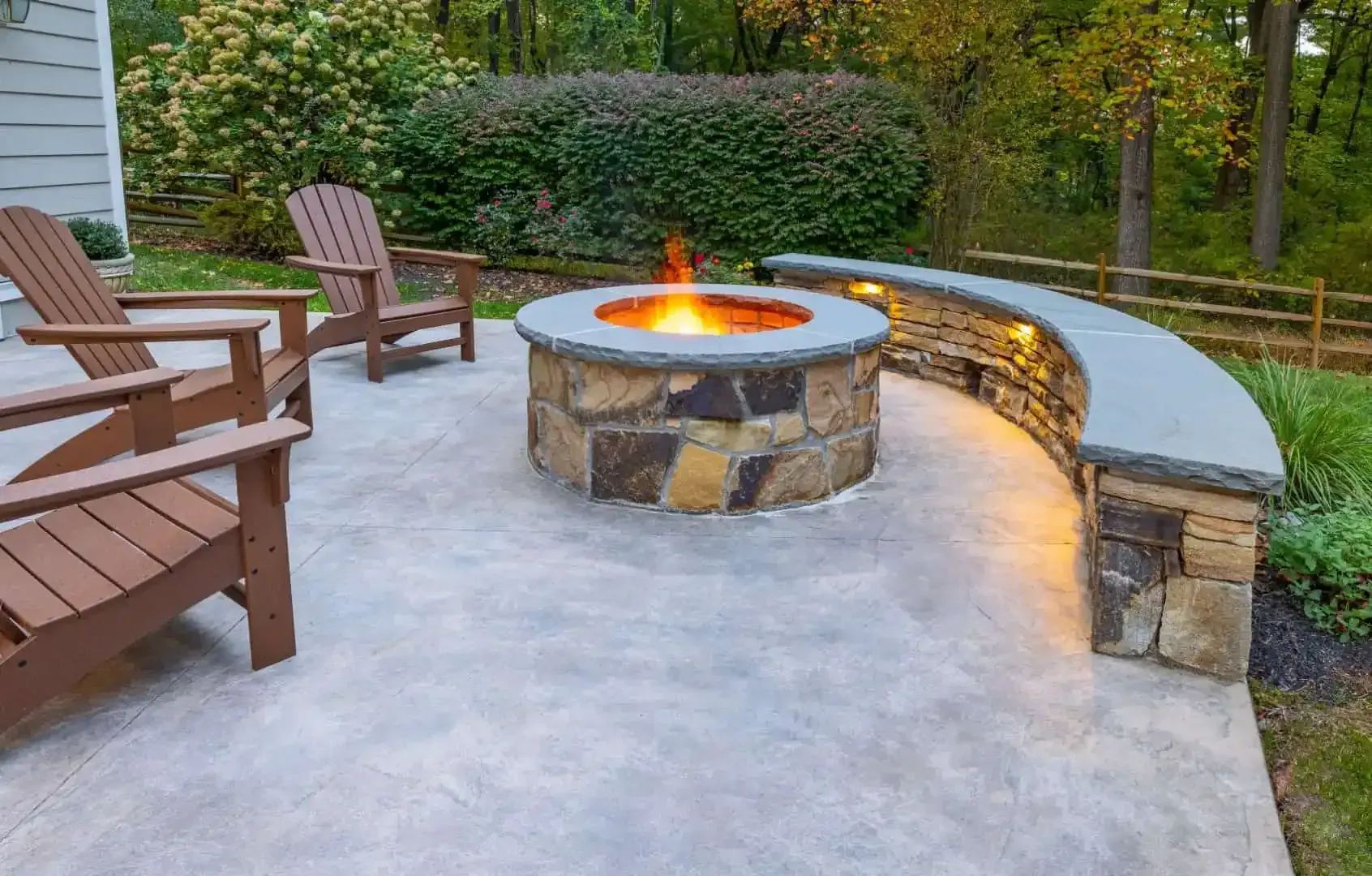 Backyard patio with stone fire pit, curved bench, and wooden chairs around a lit fire