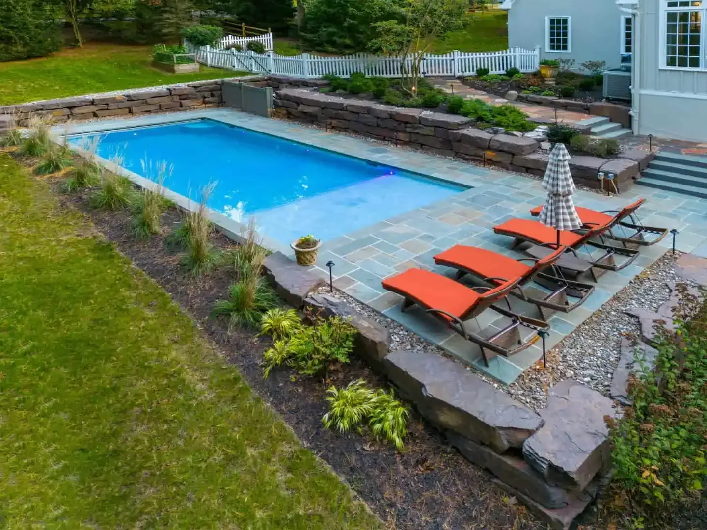 Backyard pool with orange lounge chairs, umbrella, and stone patio beside a lawn