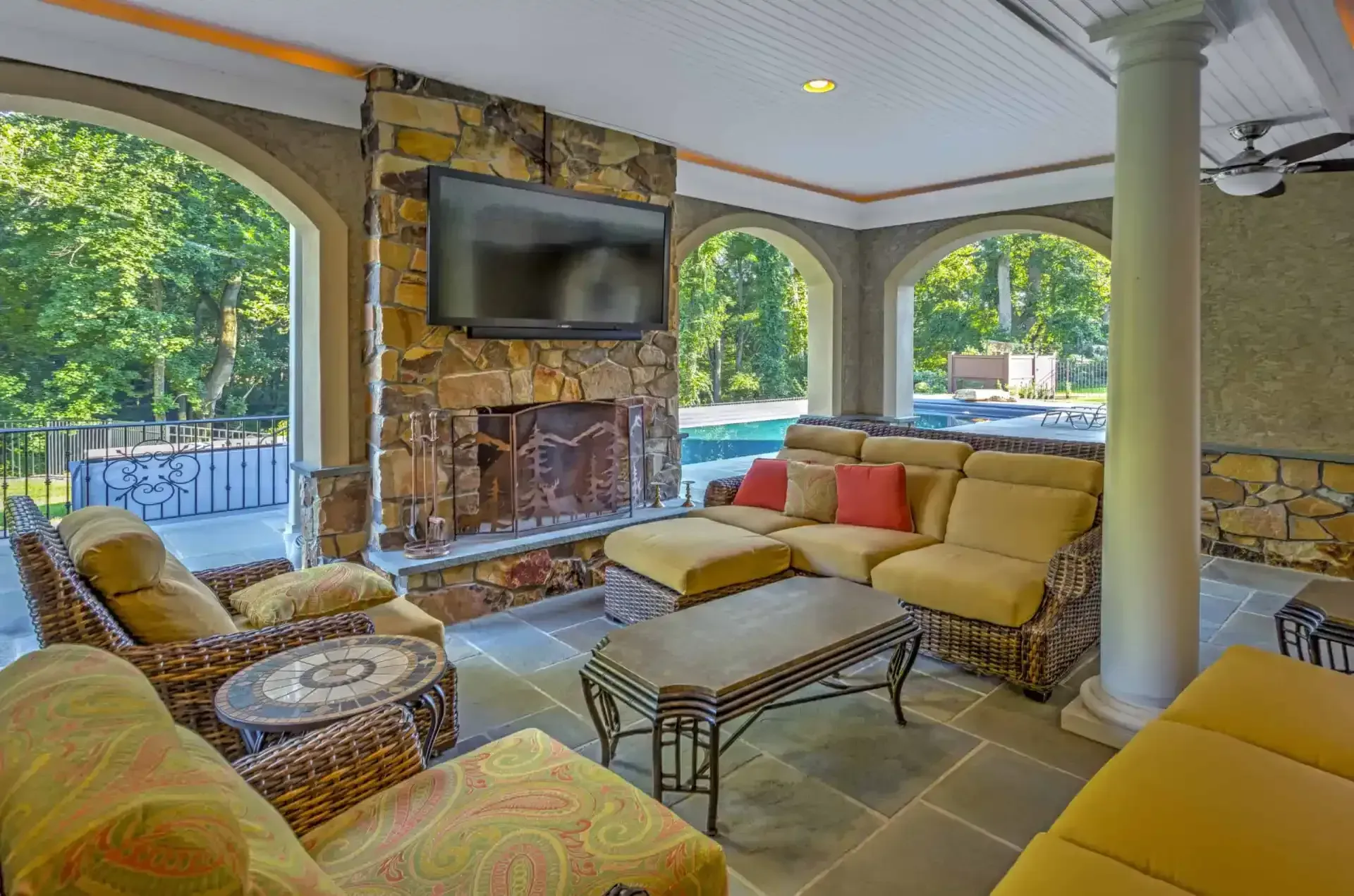 Covered patio lounge with wicker seating, fireplace, TV, and views of greenery outside