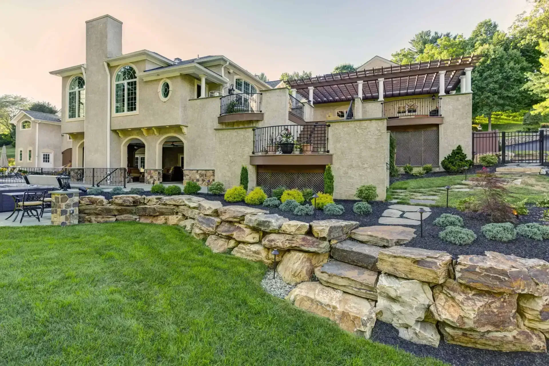Large stone house with manicured lawn, patio seating, and landscaped retaining wall in a sunny backyard