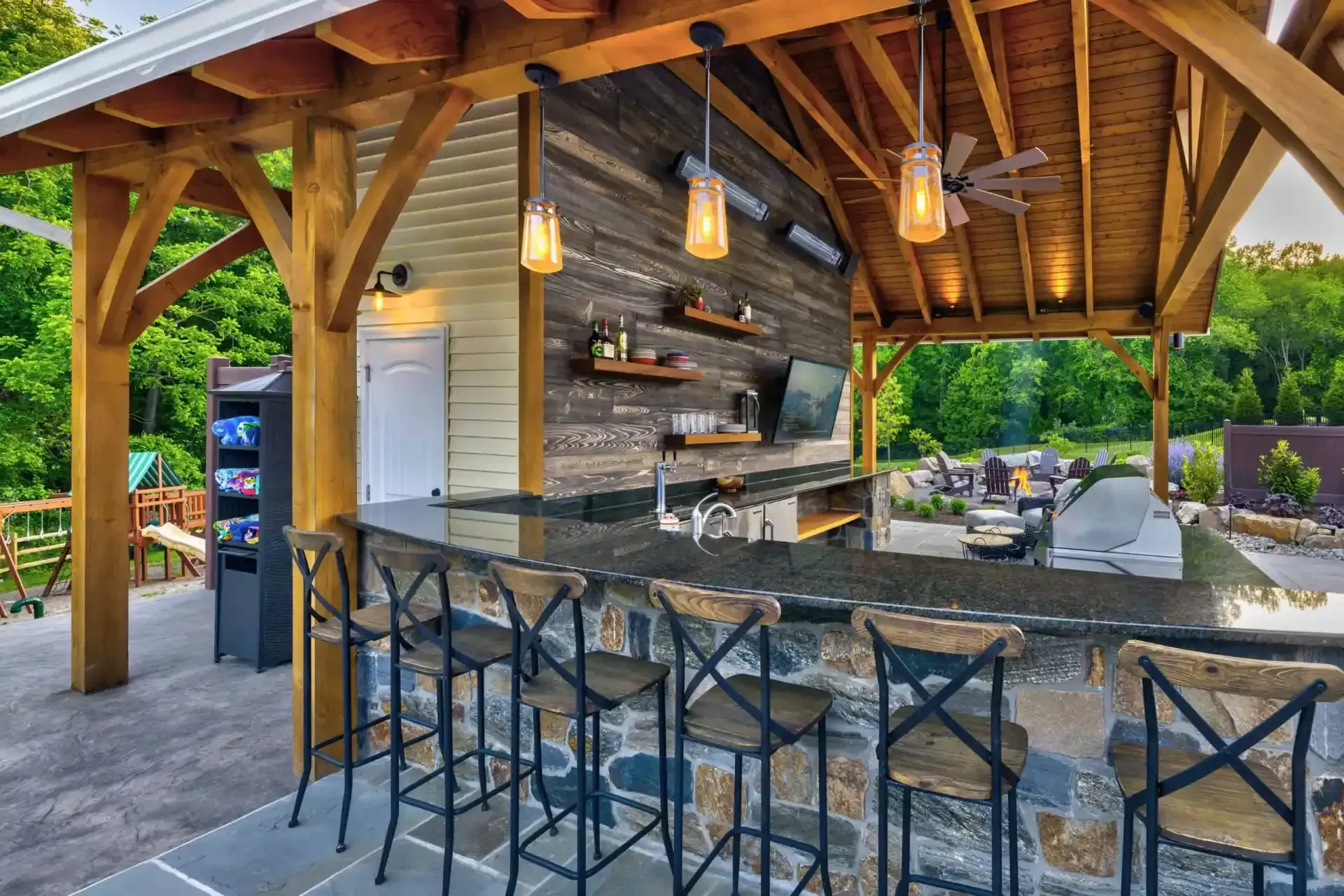 Covered outdoor kitchen and bar with stone counter, black stools, and hanging pendant lights under a wood pavilion