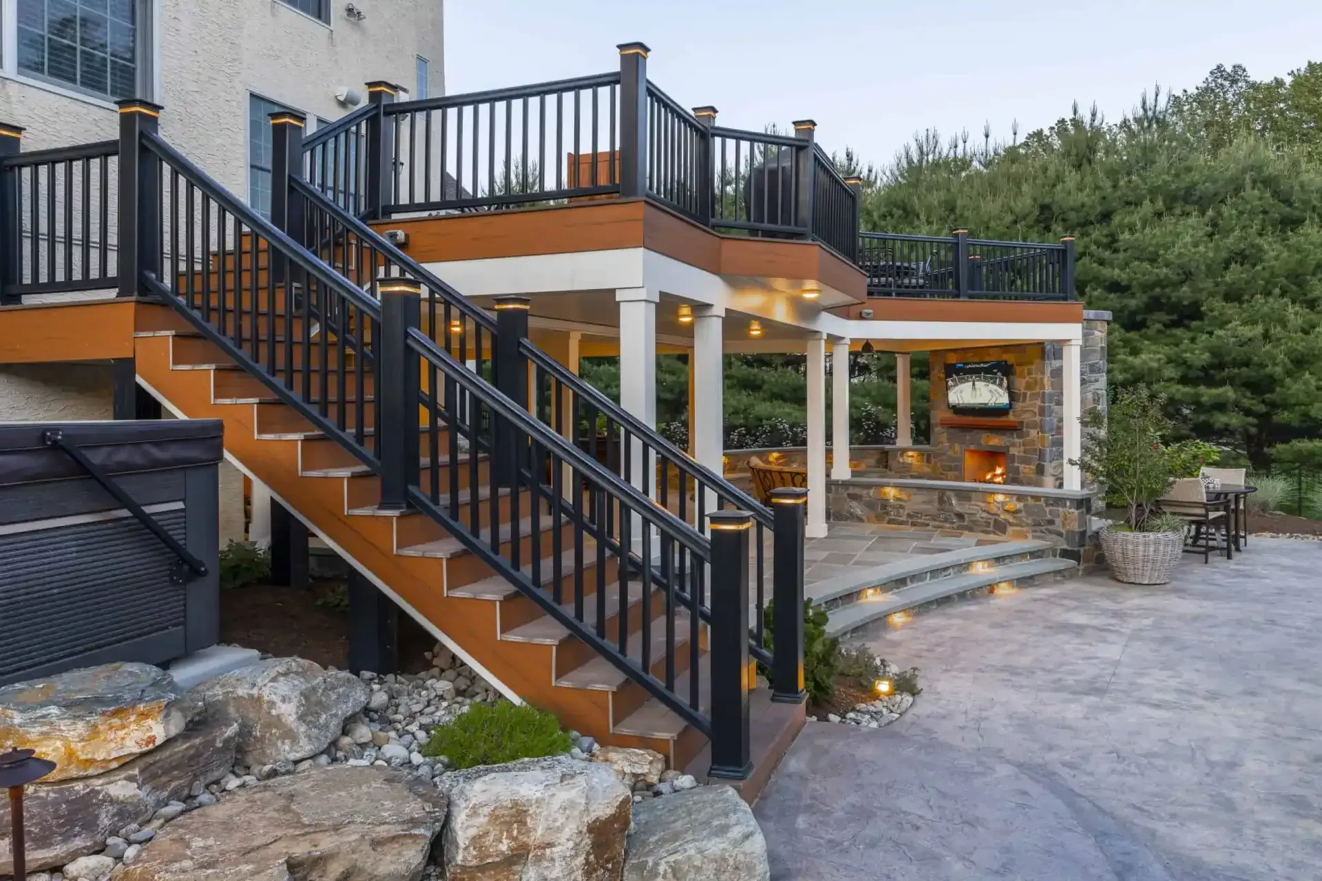 Outdoor stone patio with wooden stairs and black railings beside a house, lit by small landscape lights