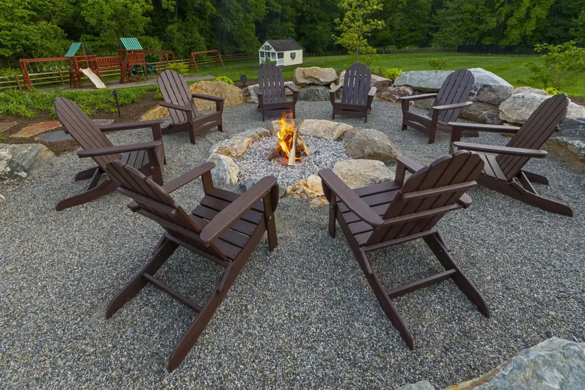 Outdoor fire pit surrounded by brown Adirondack chairs on a gravel patio, with trees and a small white house nearby