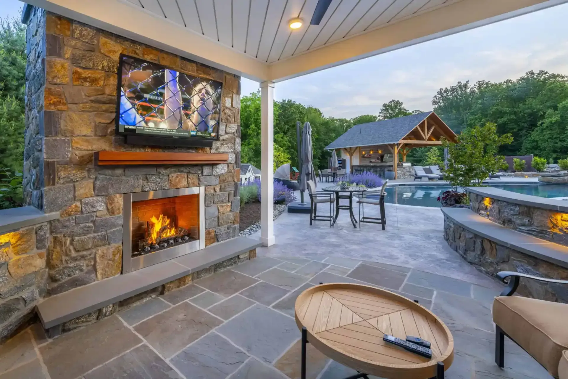 Covered patio with stone fireplace and TV, seating area, and backyard pool at dusk