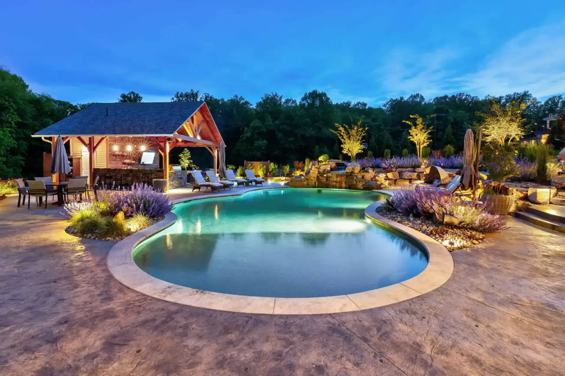 Luxury backyard pool at dusk beside a lit pavilion, surrounded by landscaped gardens and trees.