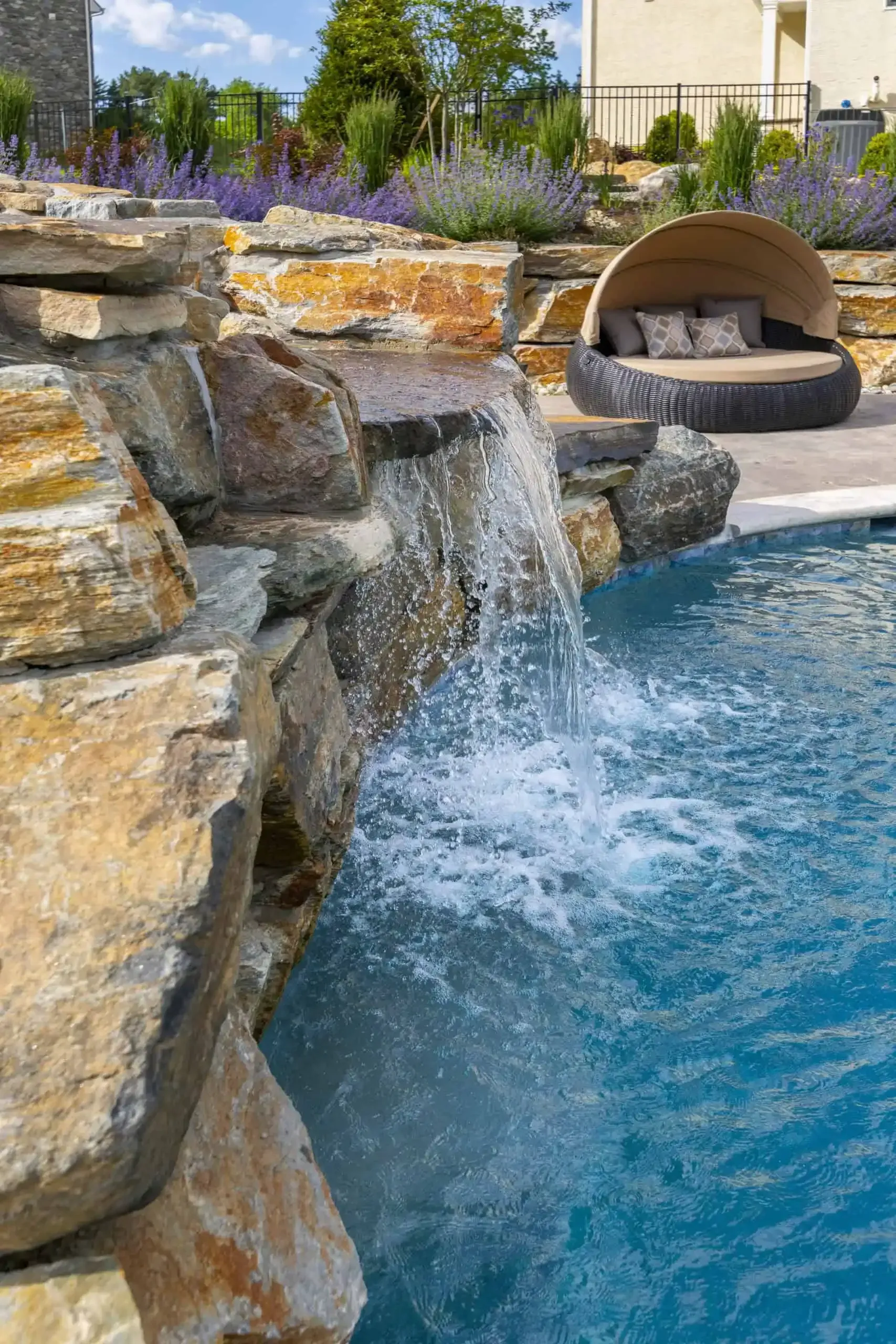 Rock waterfall cascading into a blue pool beside a stone lounge nook and landscaped garden
