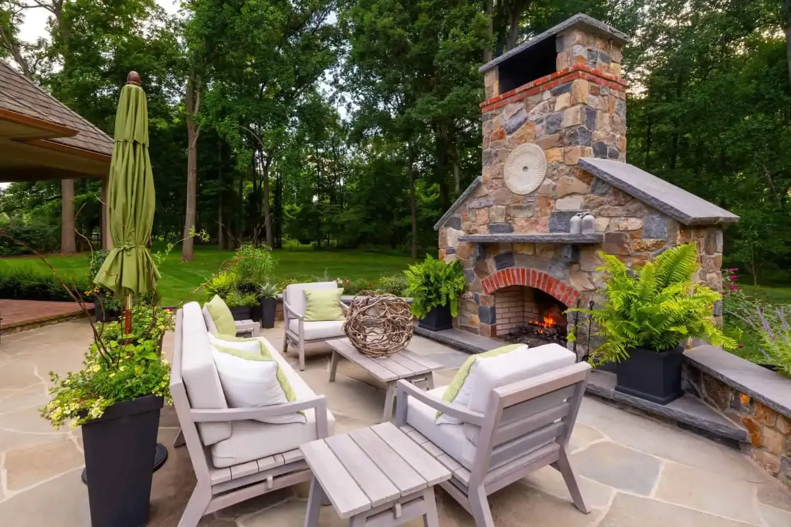 Stone patio with cushioned seating and an outdoor fireplace in a lush garden setting