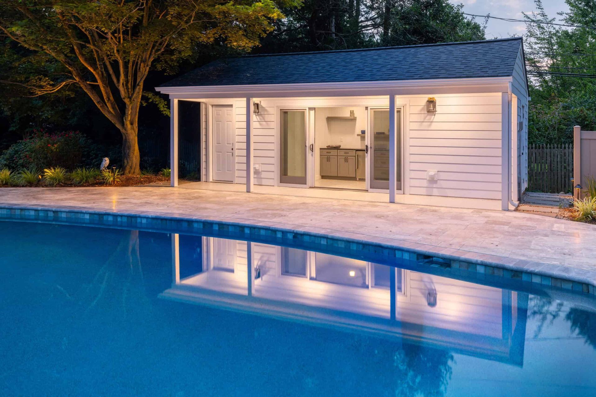 Lit white pool house at dusk reflected in a blue swimming pool