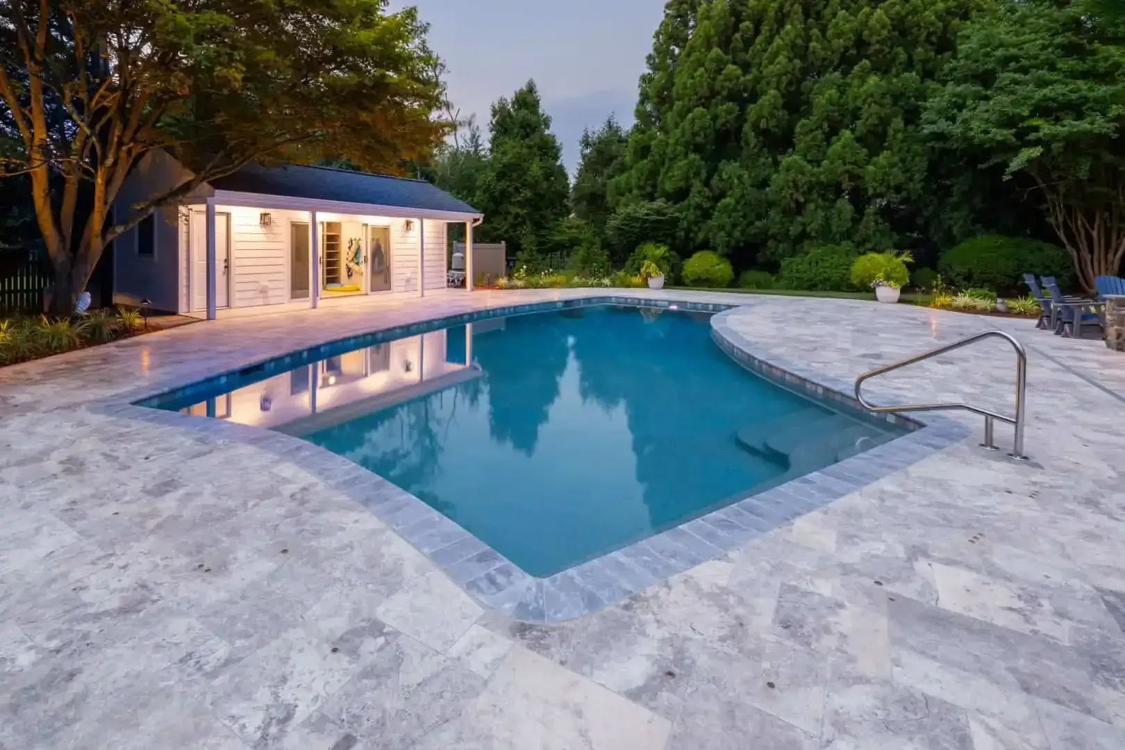 Modern backyard pool with marble deck, surrounded by trees and a lit patio at dusk