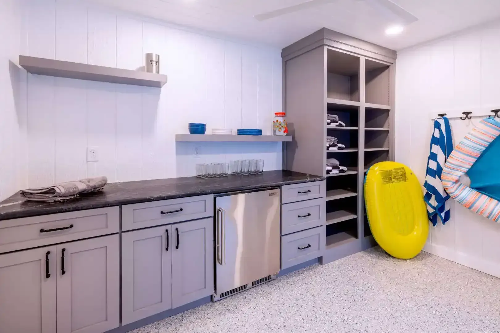 Bright utility room with gray cabinets, open shelving, stacked linens, and a yellow rescue board against the wall.