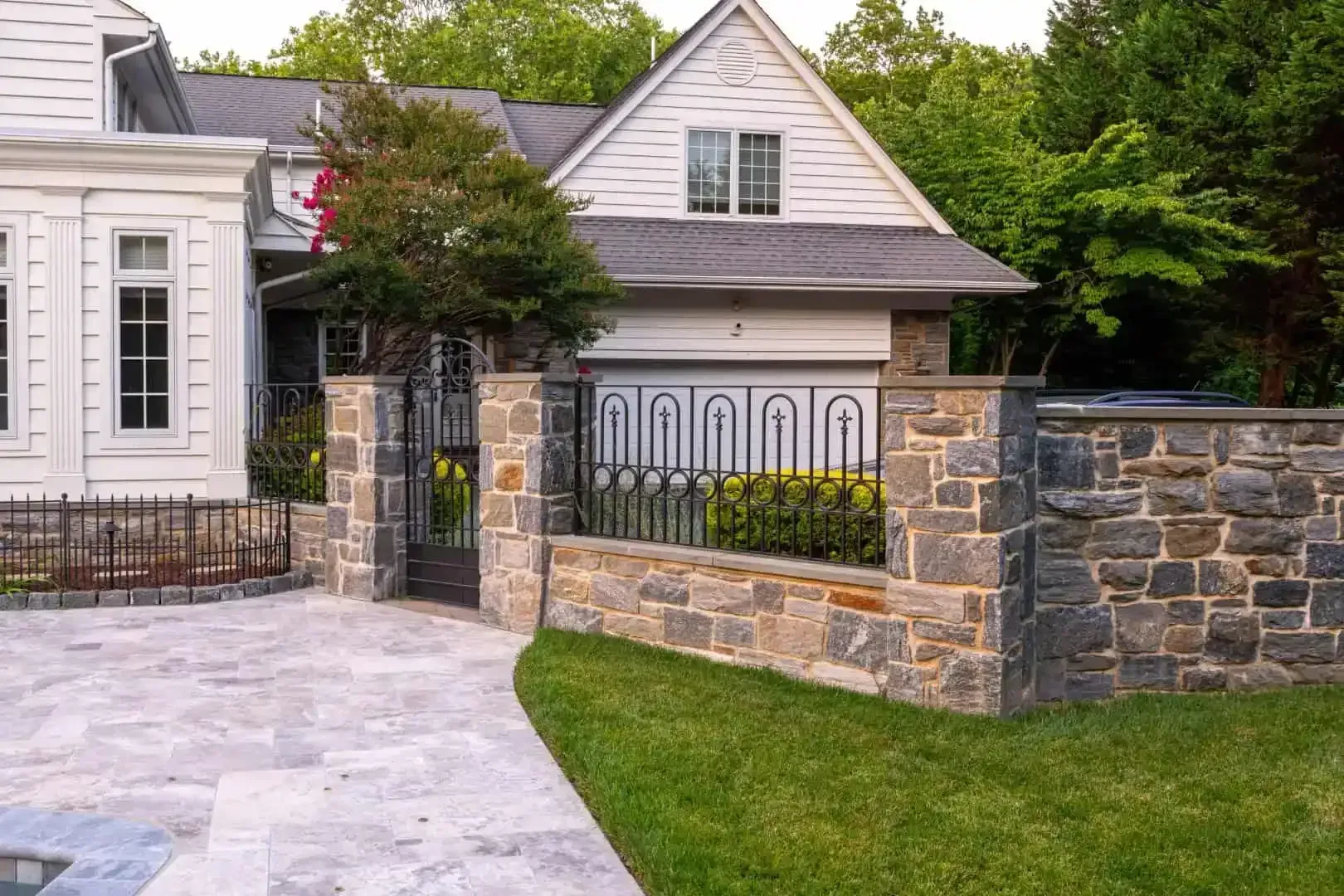 Stone house with a landscaped front yard, iron gate, and low stone wall along a paved driveway