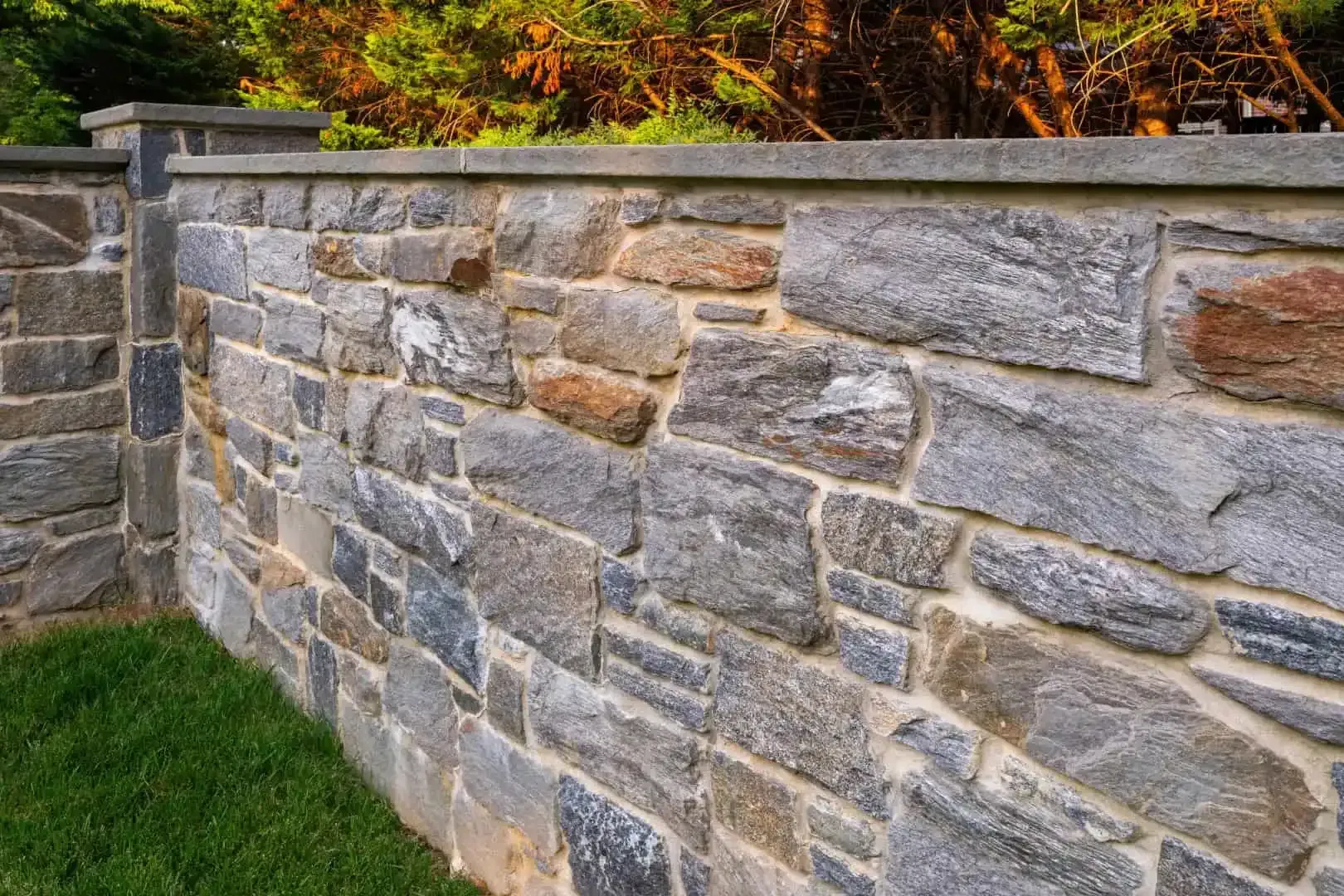 Stone retaining wall with gray and tan blocks beside green grass and autumn foliage