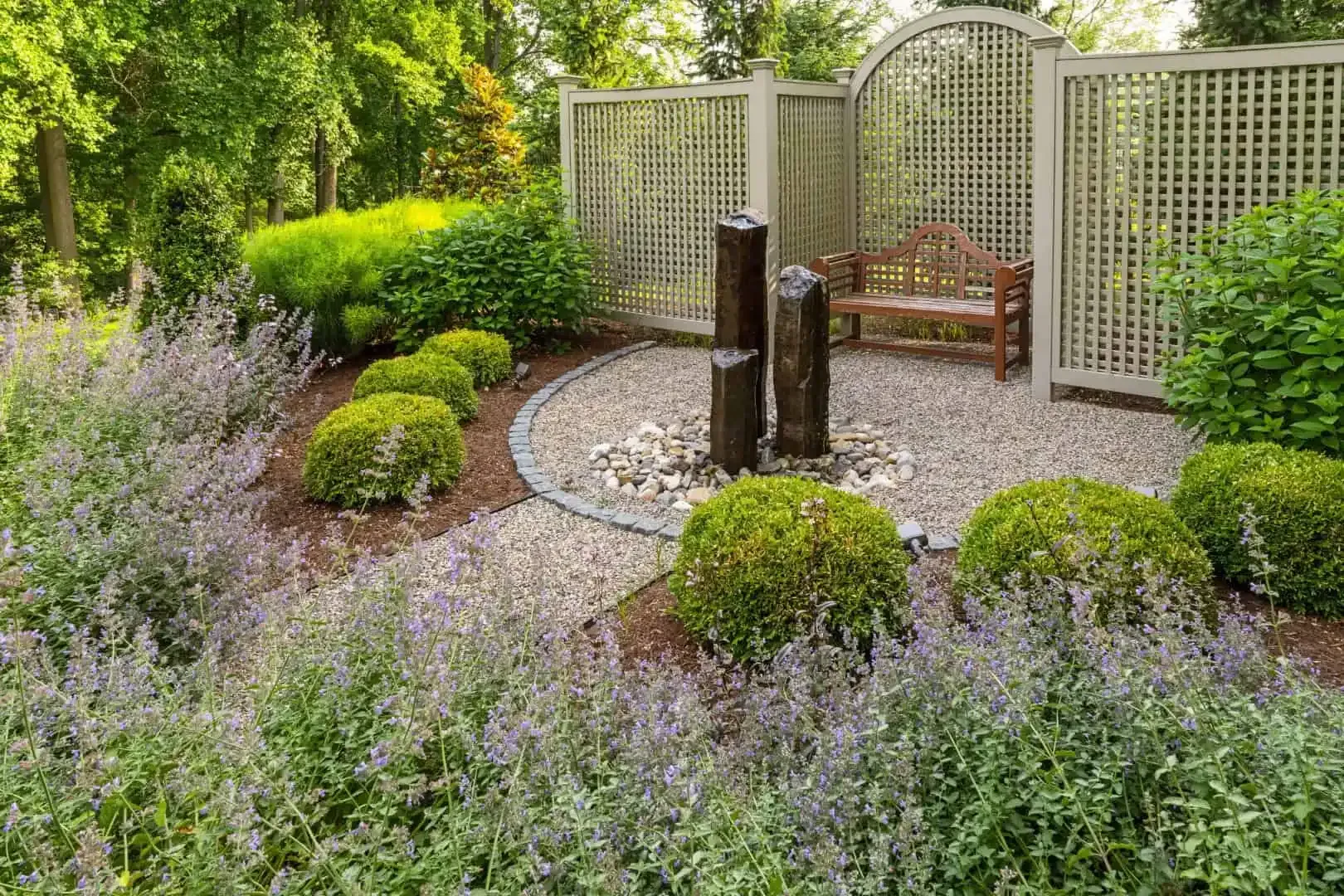 A lush garden patio with a small fountain, curved gravel path, trellis screens, and blooming lavender.
