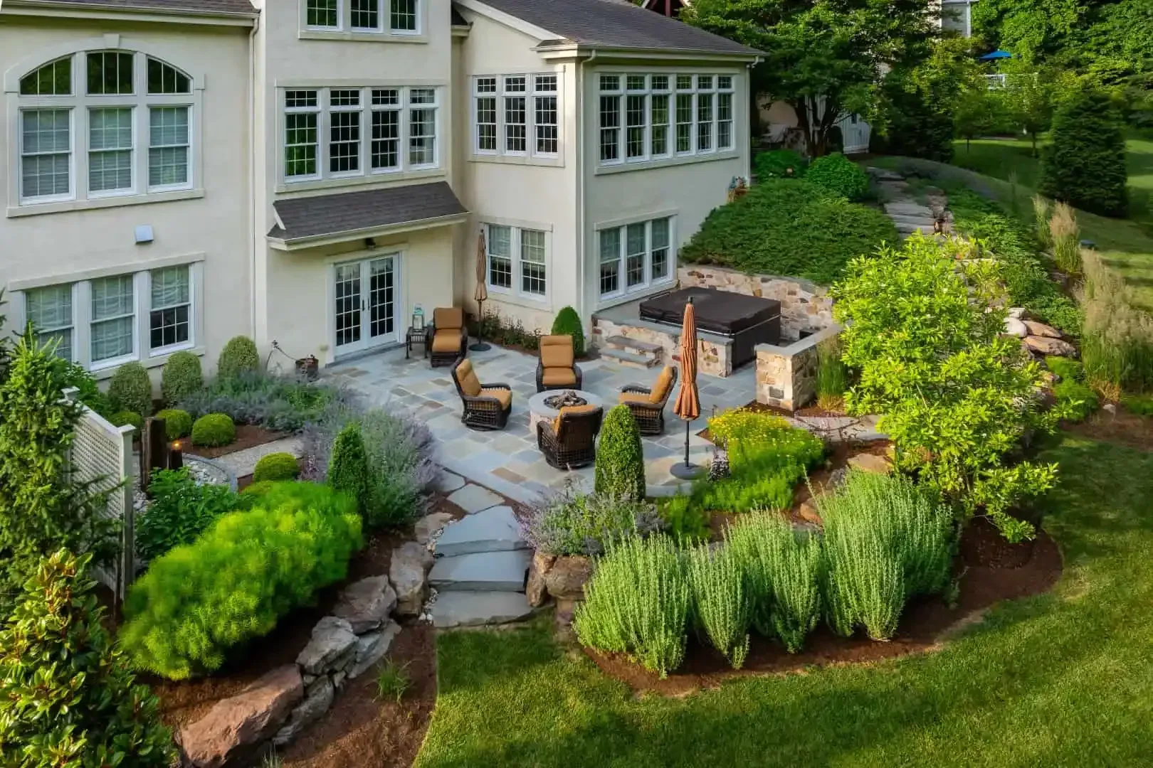 Backyard patio with outdoor seating, stone fire pit, and lush landscaping beside a white house